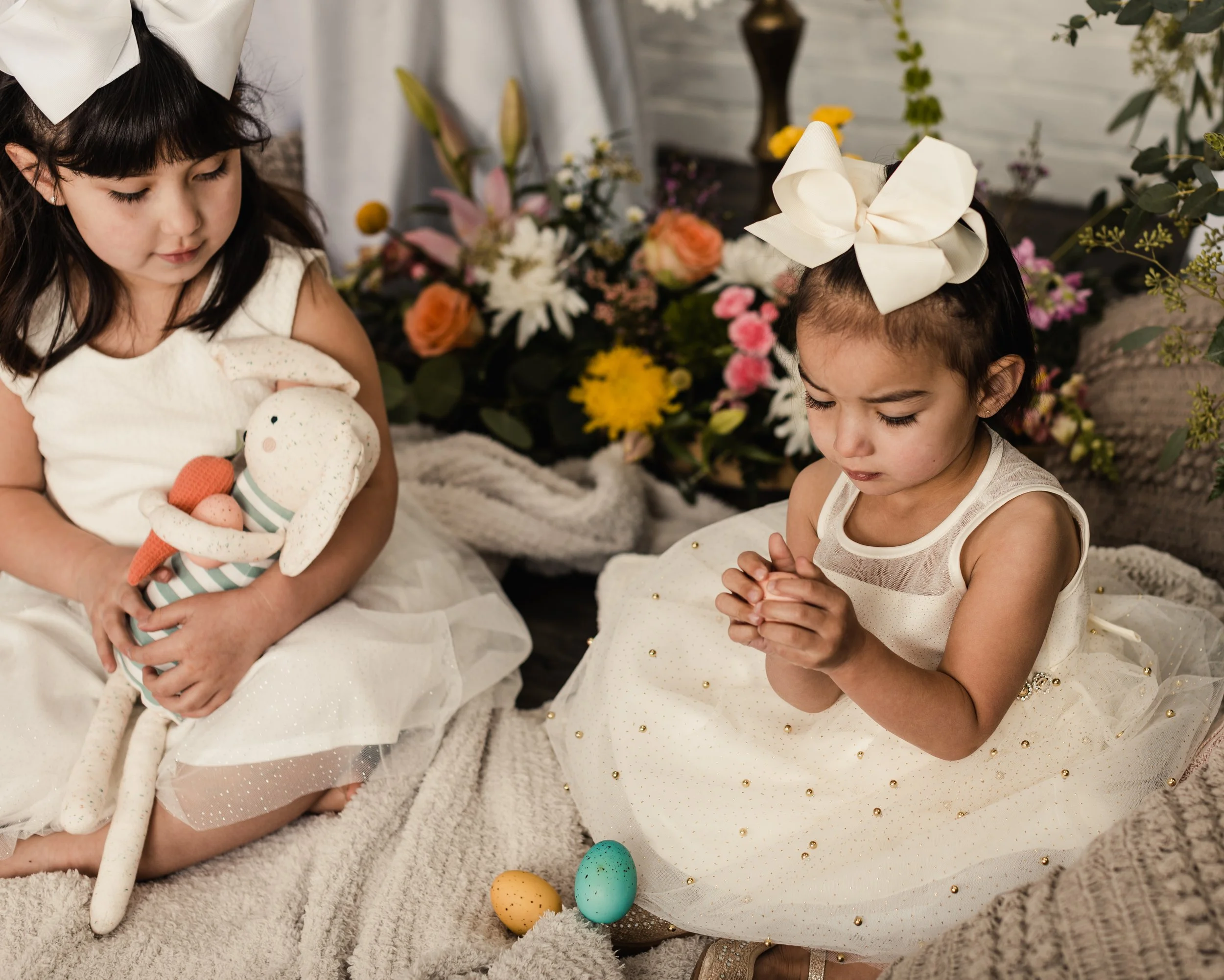 Two young girls dressed in white dresses with bows in their hair, sitting on a couch with a floral backdrop, holding decorated eggs for Easter.