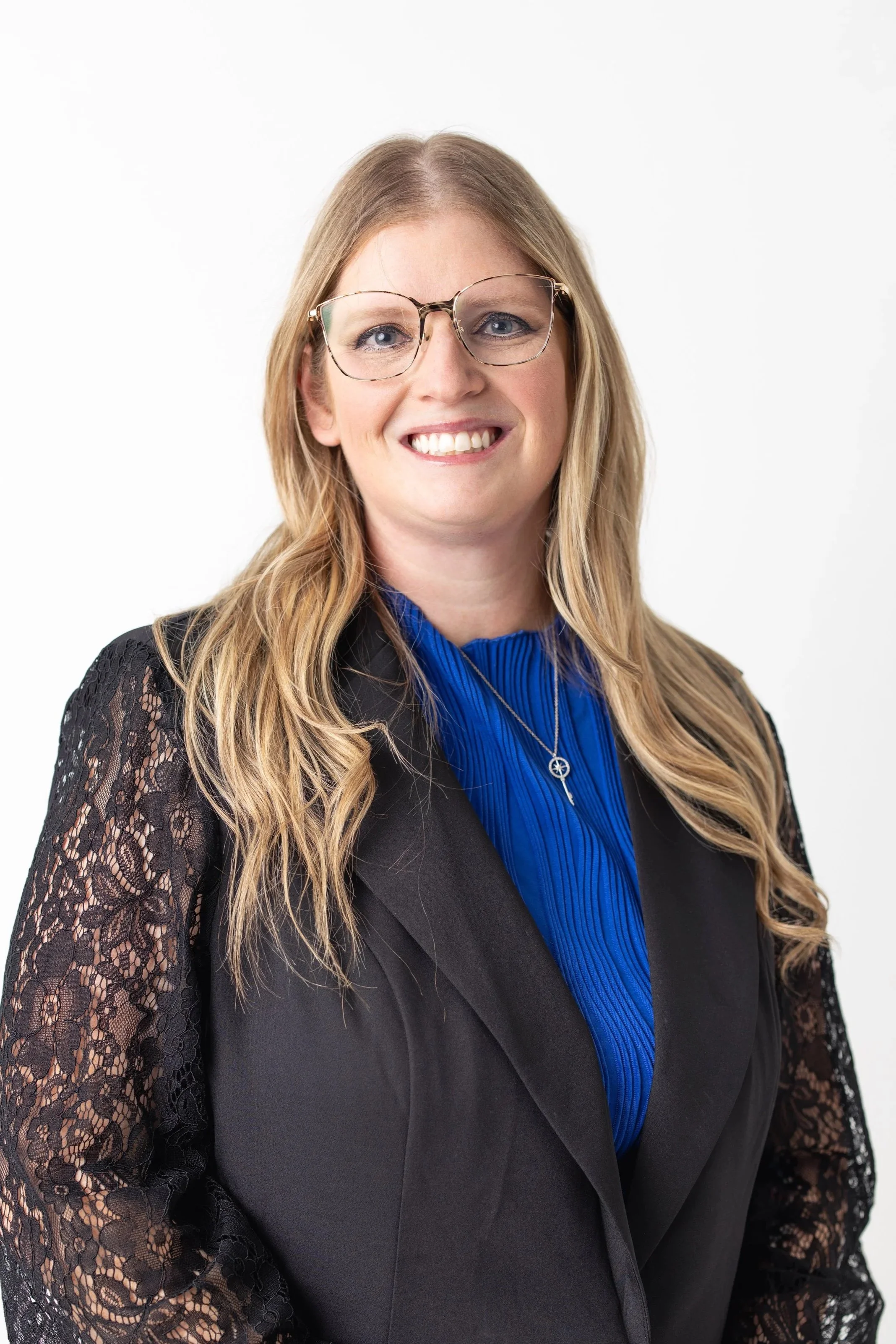 A woman with long blonde hair, wearing glasses, a black blazer with lace sleeves, a blue blouse, and a necklace with a key pendant, smiling against a plain white background.