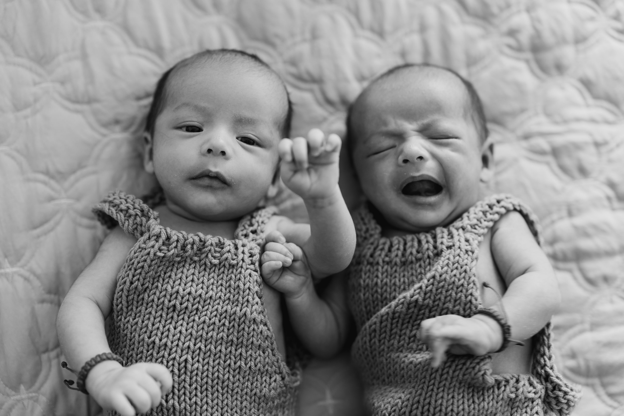 Two babies lying on a quilted blanket, one appears calm and the other is crying, both wearing knitted outfits.
