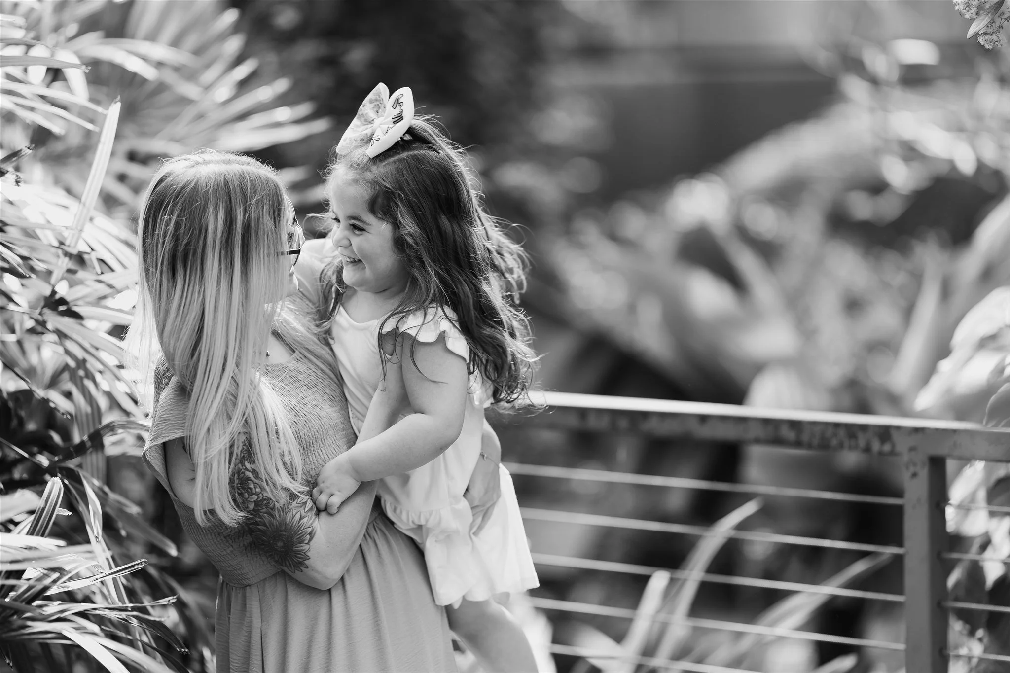 A woman holding a young girl, both smiling and looking at each other, outdoors near a railing with lush foliage in the background.