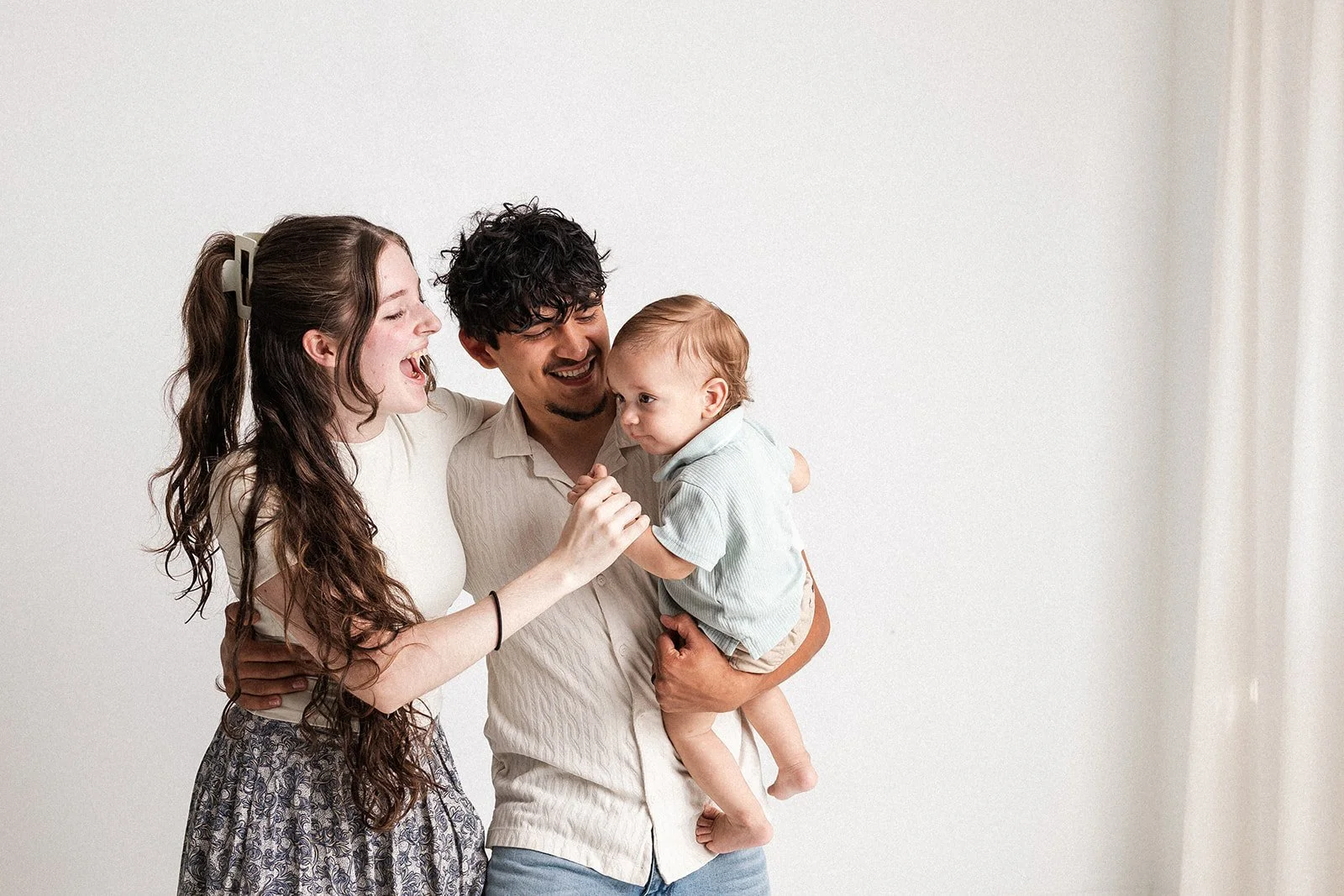 A family of three, a young woman, a man, and a small child, smiling and playing together in a bright room.