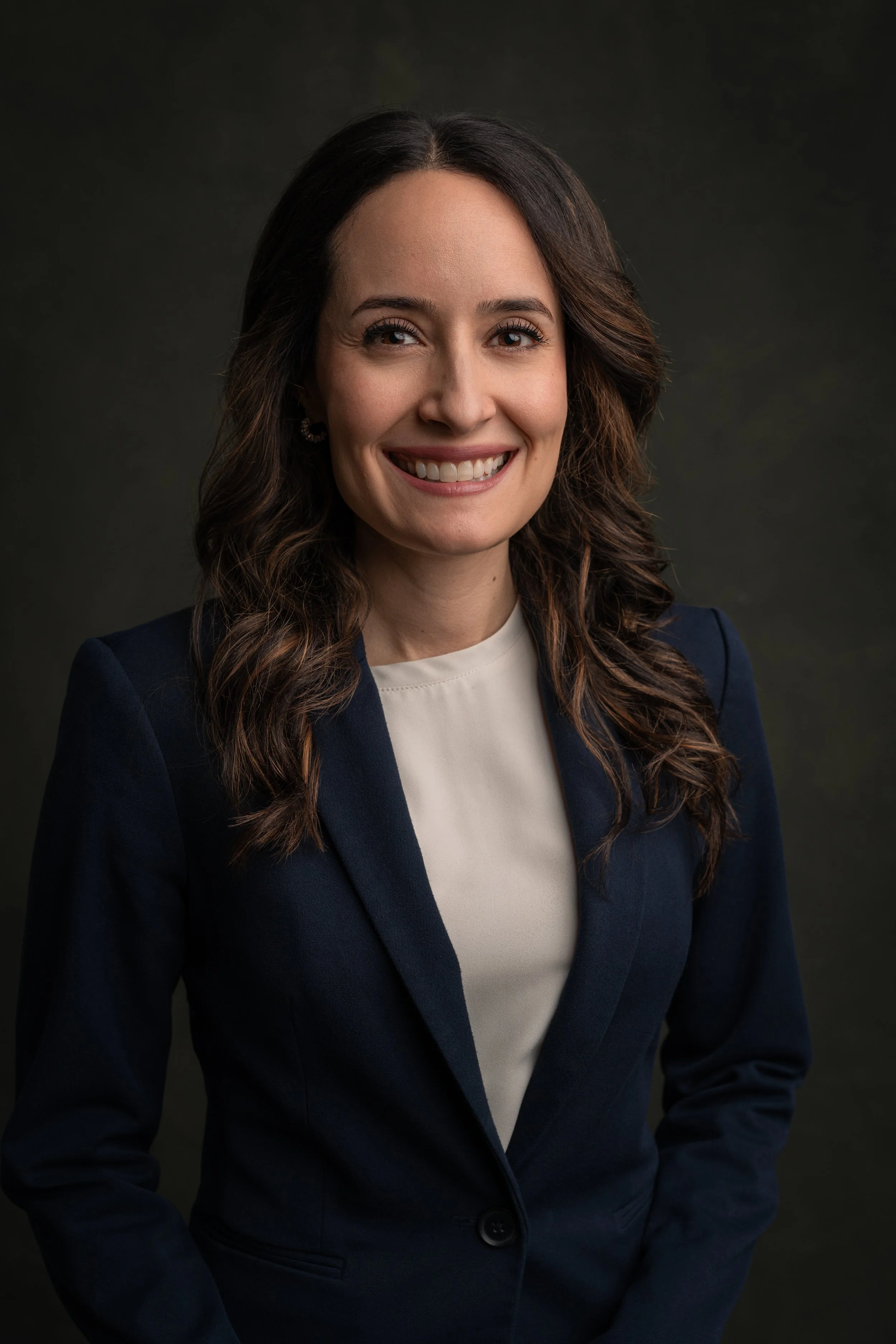 A professional woman with wavy brown hair, smiling, wearing a navy blue blazer and a beige blouse, against a dark backdrop.