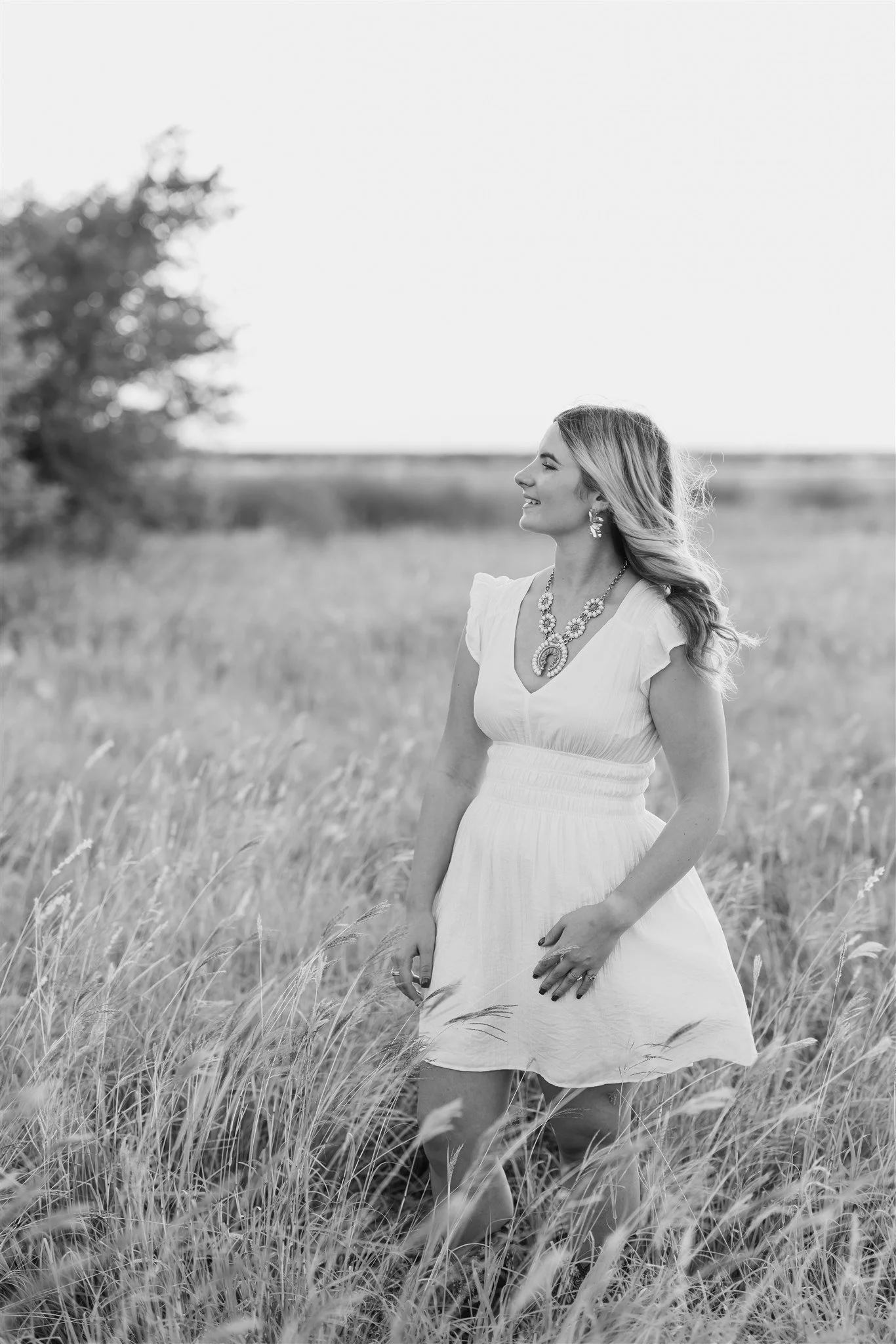 A woman in a white dress standing in a field of tall grass during daylight, smiling and looking to her right, with jewelry including a necklace and earrings. Black and white photo.