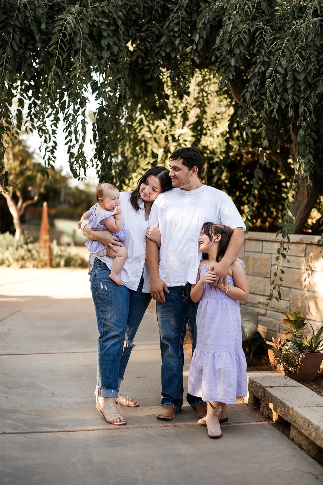 Family of four enjoying an outdoor walk beneath a large tree, smiling and holding each other.