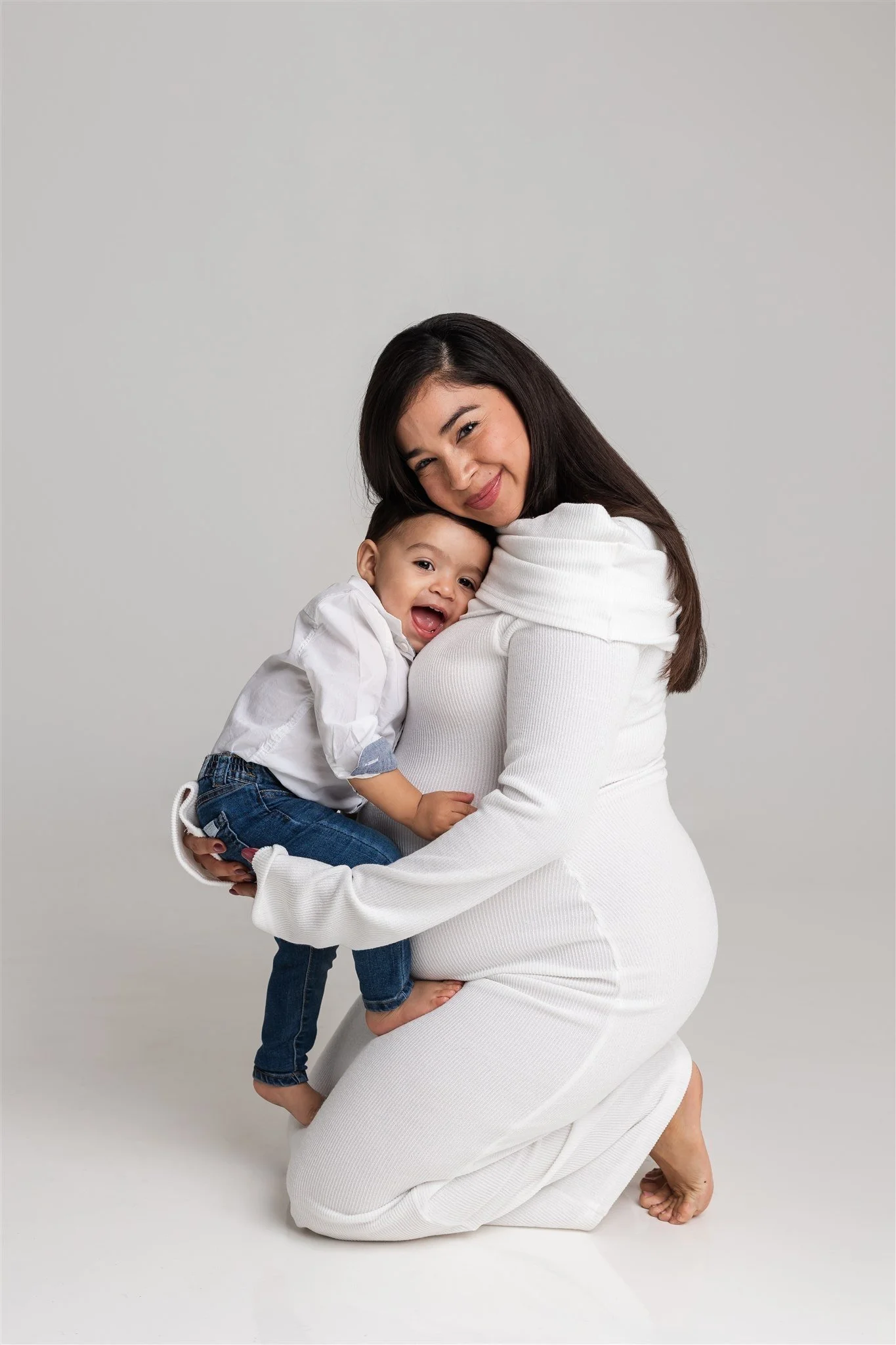 A woman with long dark hair dressed in a white outfit is kneeling and holding a smiling young child with short dark hair, wearing a white shirt and blue jeans, against a plain light grey background.