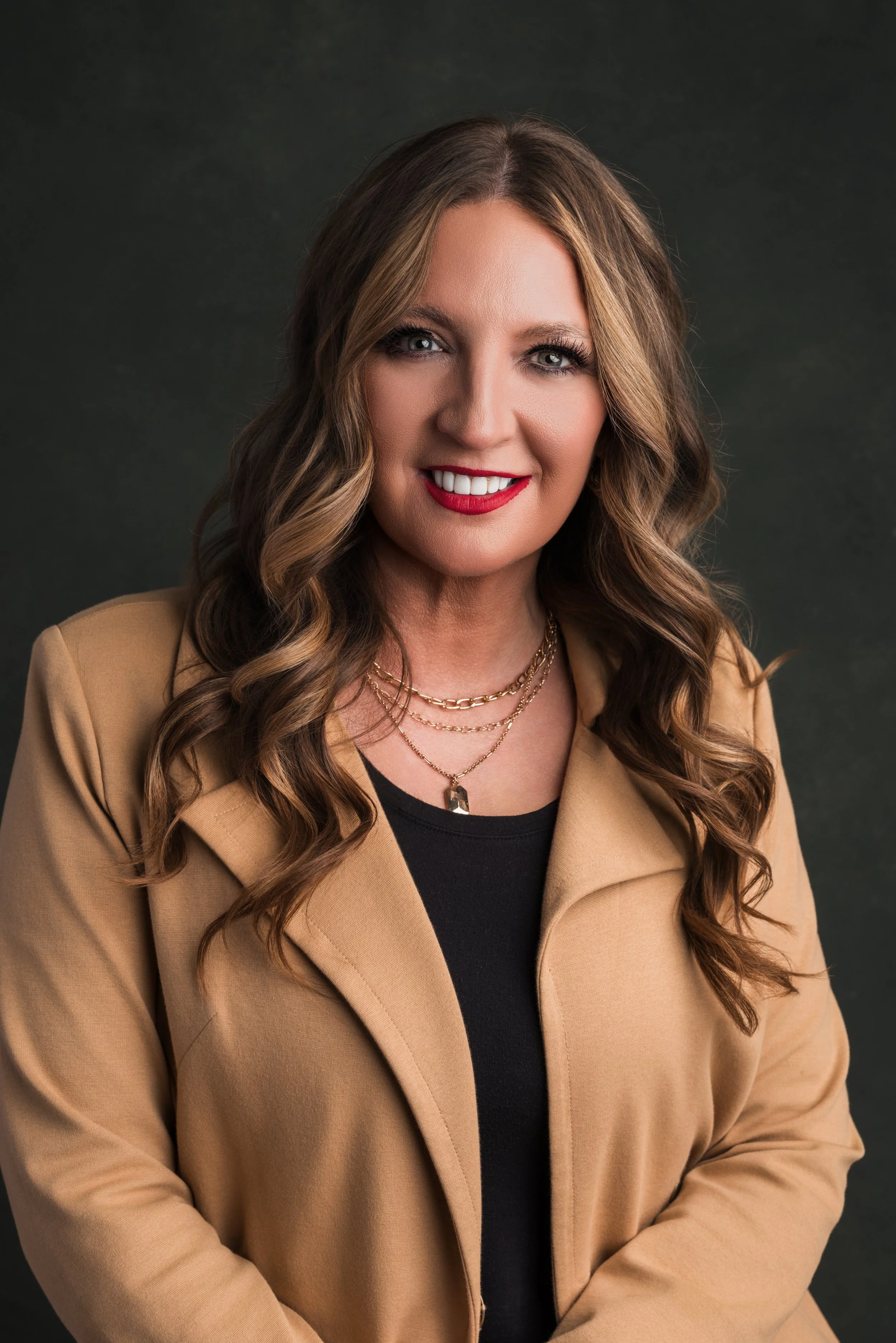 A woman with brown, wavy hair smiling, wearing a tan blazer, black top, layered gold necklaces, and red lipstick against a dark background.