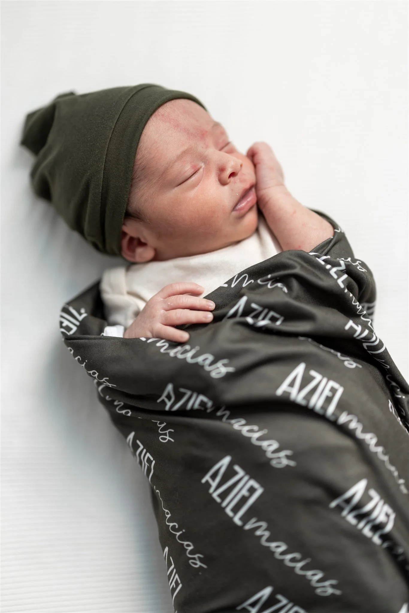 A newborn baby lying on a white surface, wearing a green hat and wrapped in a black blanket with white text, sleeping peacefully with a hand resting on the face.