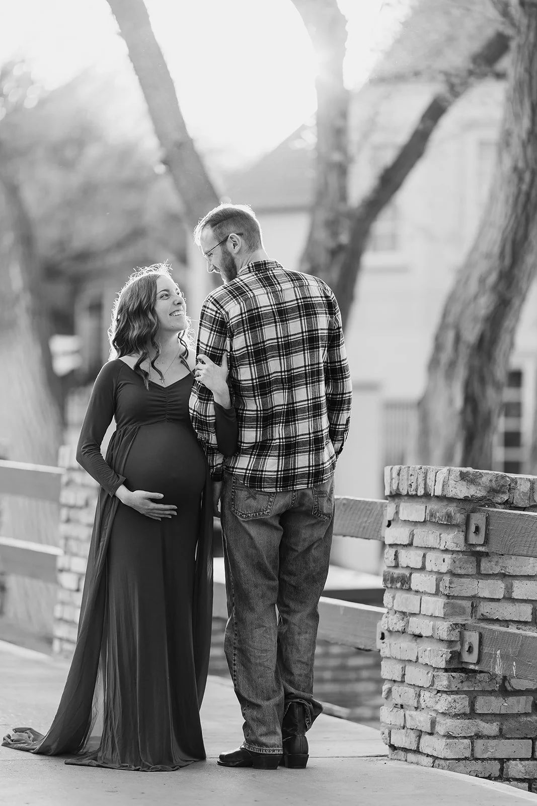 A pregnant woman and a man are standing outdoors, facing each other with smiles. The woman has long, wavy hair and is wearing a long dress, and the man has short hair, a beard, glasses, and is dressed in a plaid shirt and jeans. They are holding hand