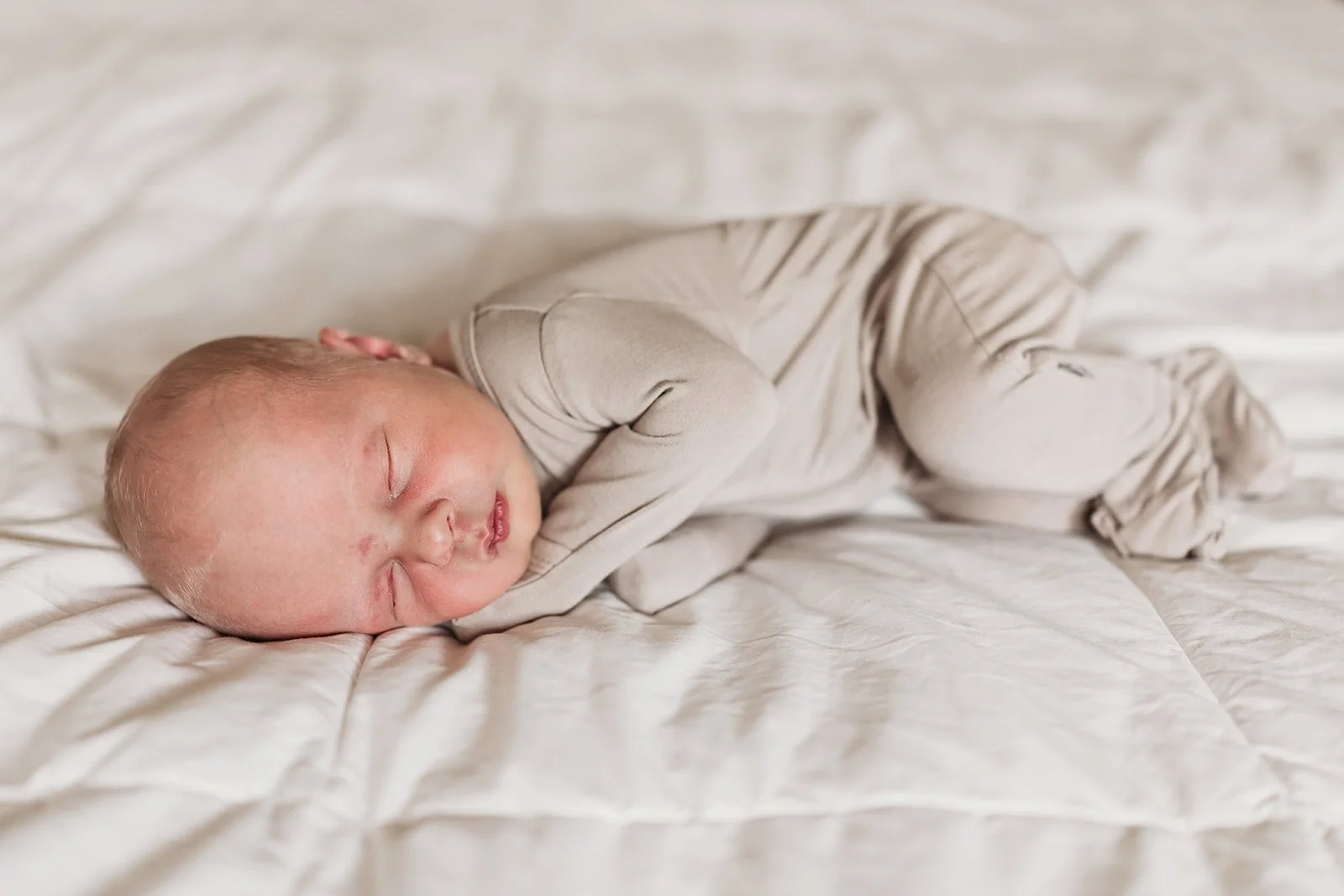 A sleeping baby lying on a white bed in beige clothing.