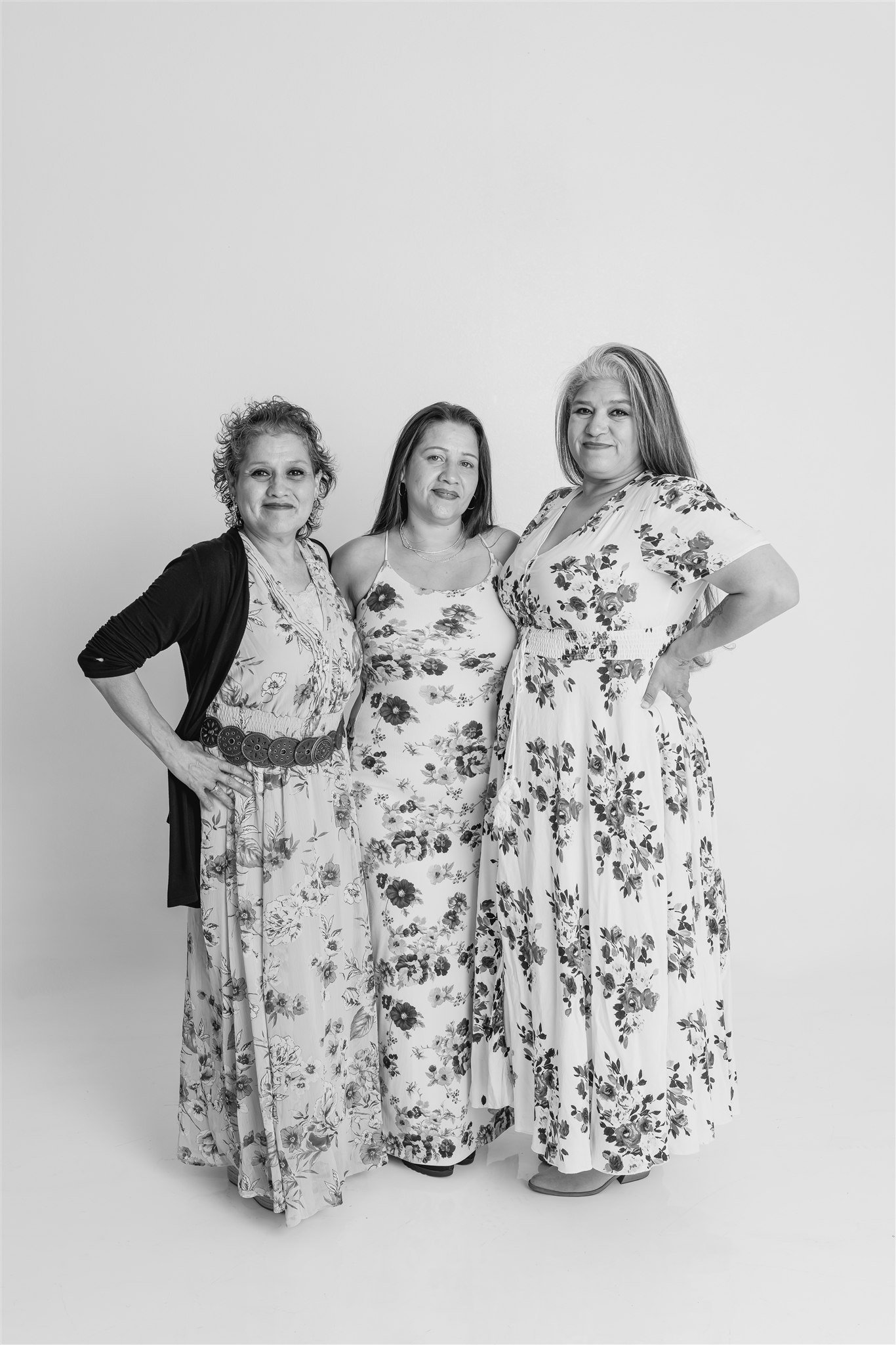 Three women standing together, dressed in floral dresses, posing for a photograph against a plain background.