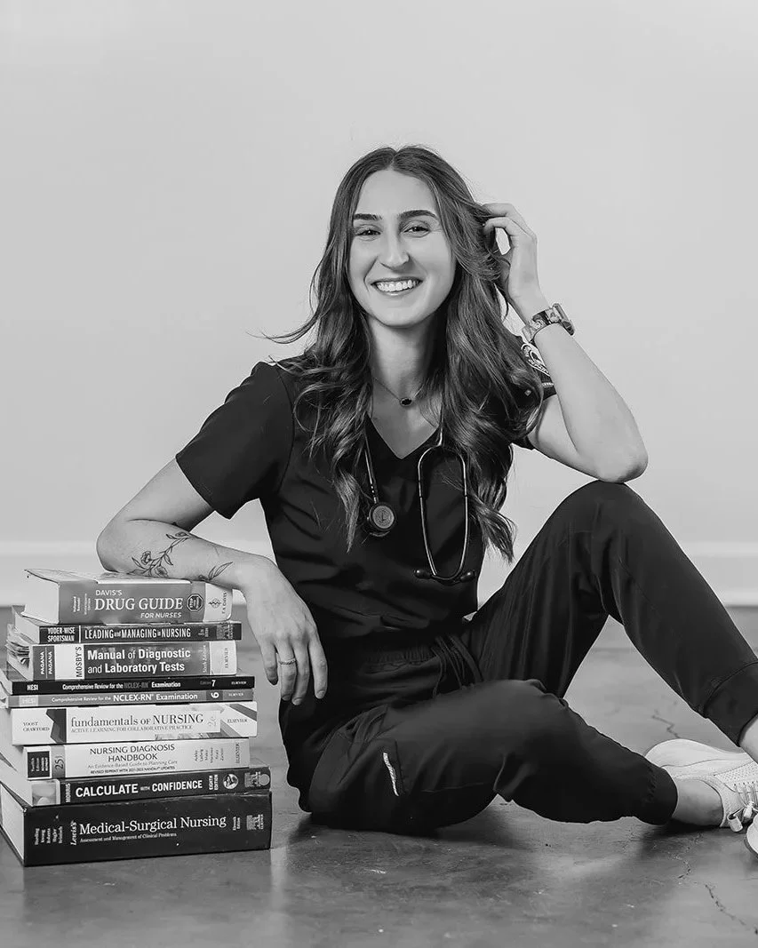 A young woman in scrubs sitting on the floor, smiling, with a stack of nursing and medical books beside her, and a stethoscope around her neck. Senior, WT Nursing student graduation photos.