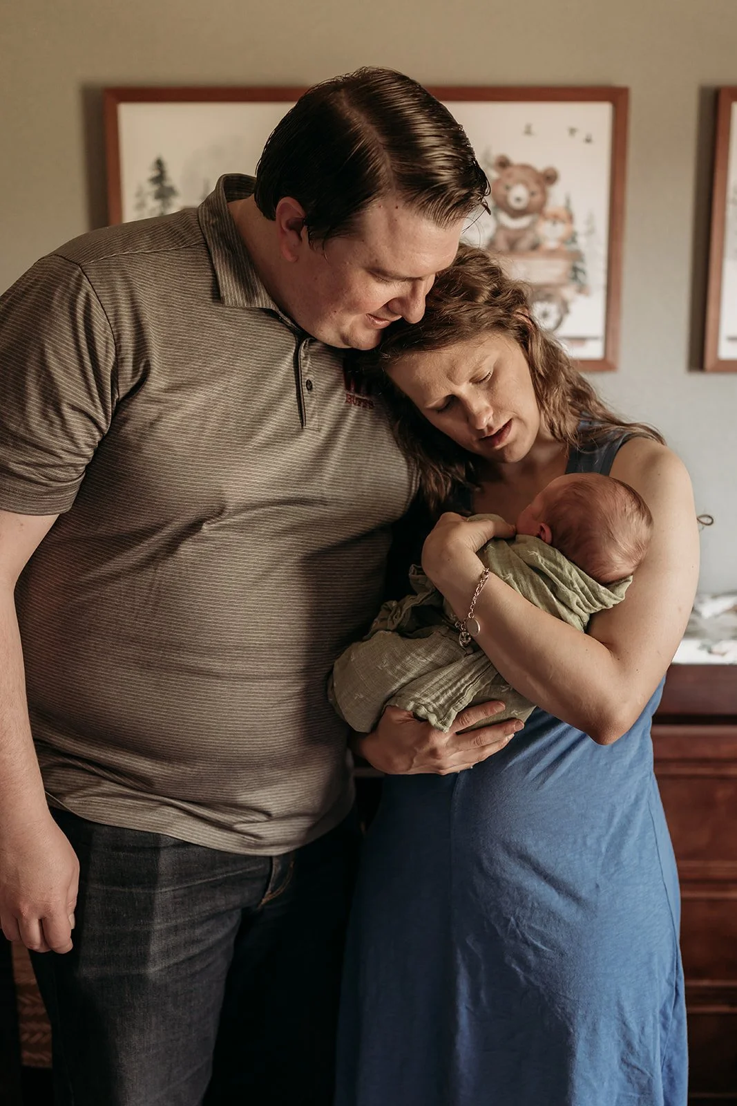 A couple holding their newborn baby in a bedroom, with framed pictures on the wall behind them. In-home newborn session near Canyon, Texas, focuses on emotion and candid moments.