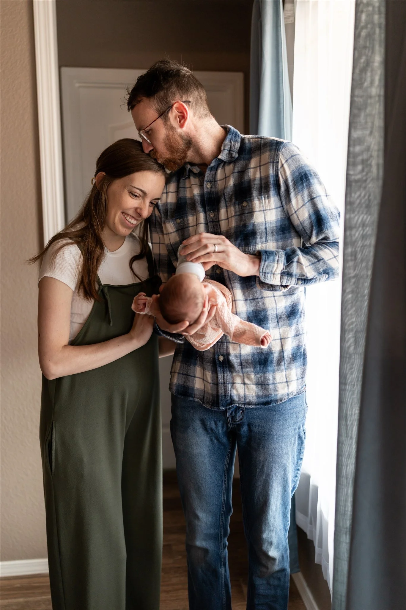 A couple holding a newborn baby in a cozy room with natural light. The woman is smiling at the baby, and the man is gently feeding the baby with a bottle.