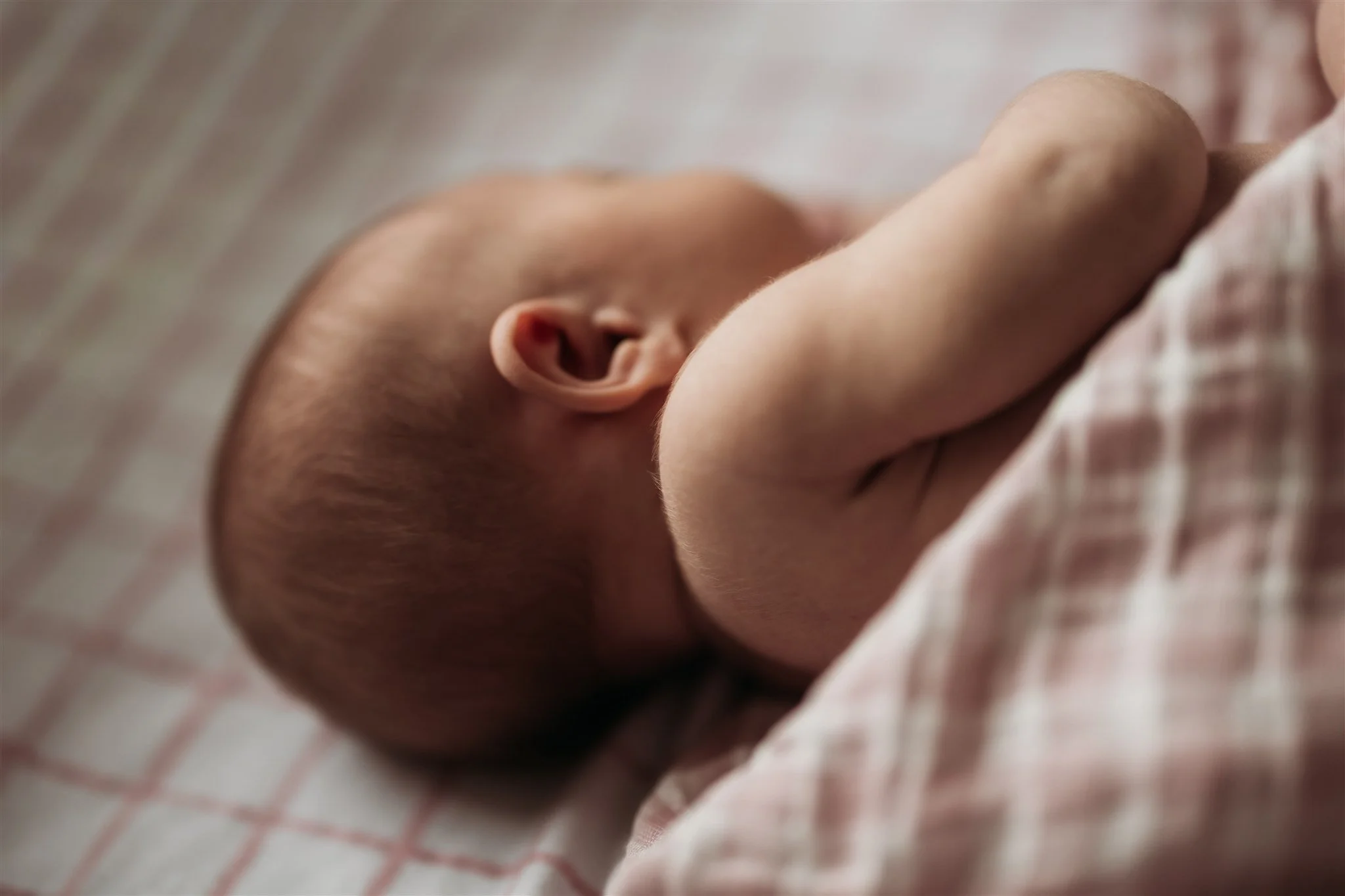 A close-up of a sleeping baby lying on a checkered blanket, showing the side of the baby's face, ear, and arm.