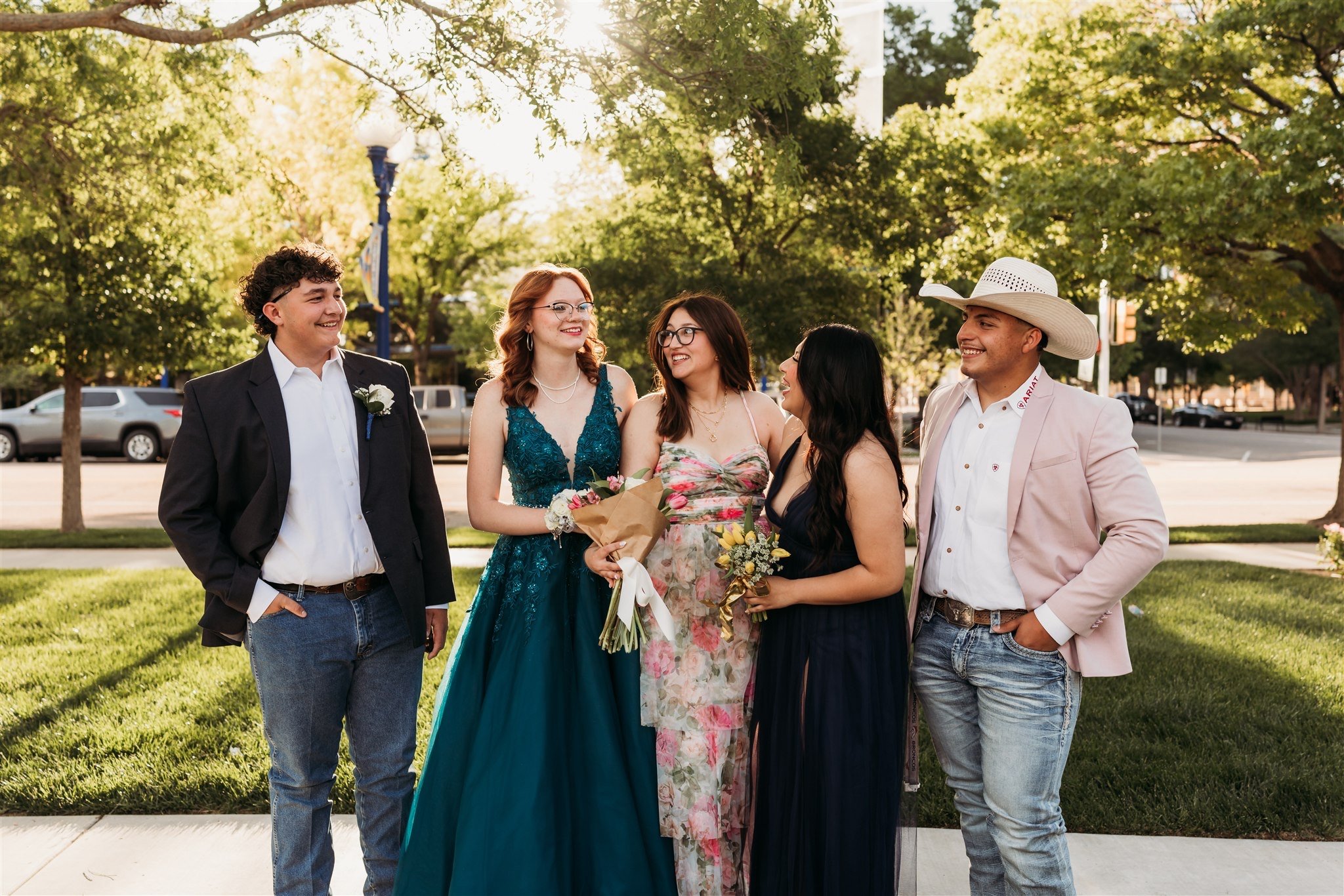 A group of five people standing outdoors on a sunny day, dressed in formal and semi-formal attire, with trees and parked cars in the background. They are smiling and conversing.