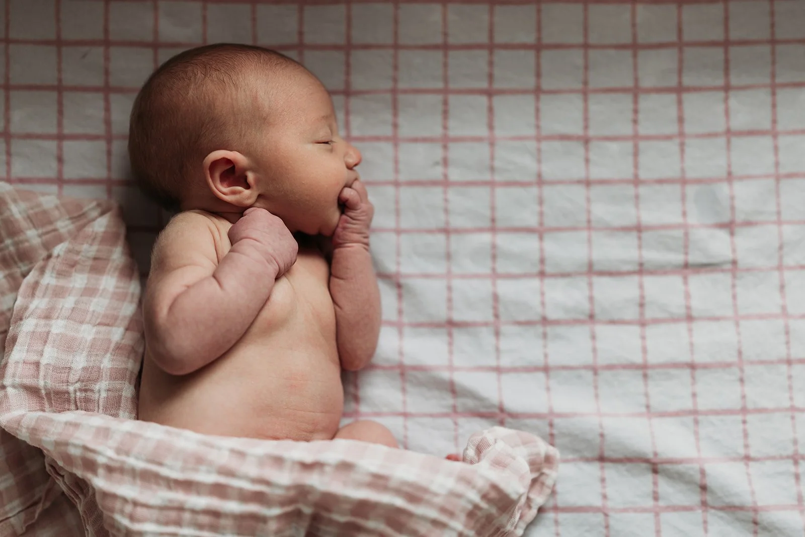 A sleeping newborn baby lying on a checkered pink and white blanket, with one hand near its face and the other bent at the elbow. An in-home newborn session near Canyon, Texas, focuses on emotion and candid moments.