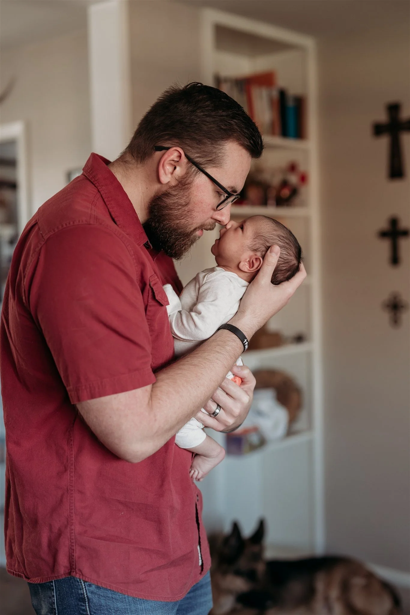 A man with glasses and a beard holding a newborn baby close to his face indoors.