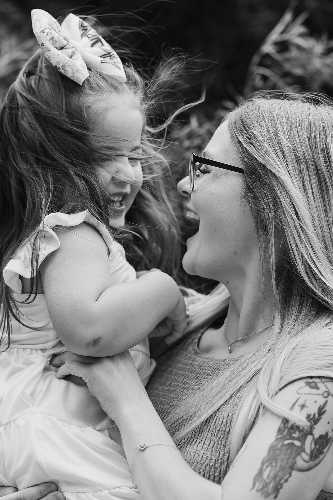 A woman with glasses smiling and holding a young girl with long hair and a bow, both joyfully interacting with their faces close together outdoors.
