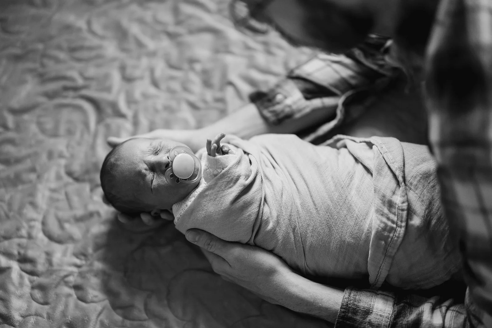 A newborn baby with a pacifier lying on a textured blanket, being held gently by an adult dad in a plaid shirt. In-home session near amarillo, texas.