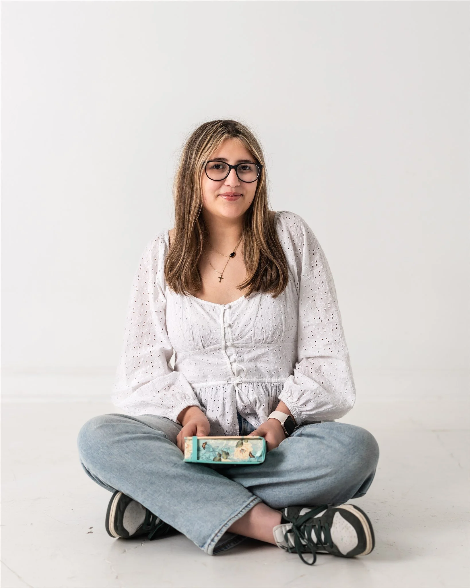 A young woman with long brown hair, glasses, and gold jewelry sits cross-legged on the floor against a plain white background, holding a small book or notebook with a floral cover.