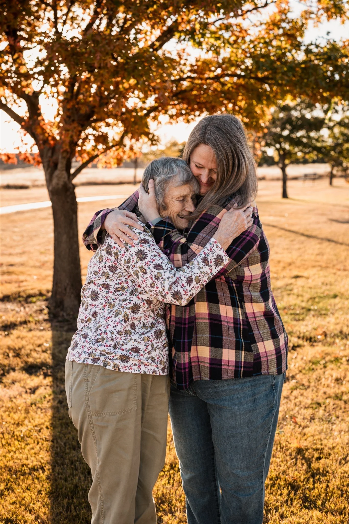 A young woman and elderly woman hugging outdoors during autumn with orange and yellow leaves, expressing joy and affection.