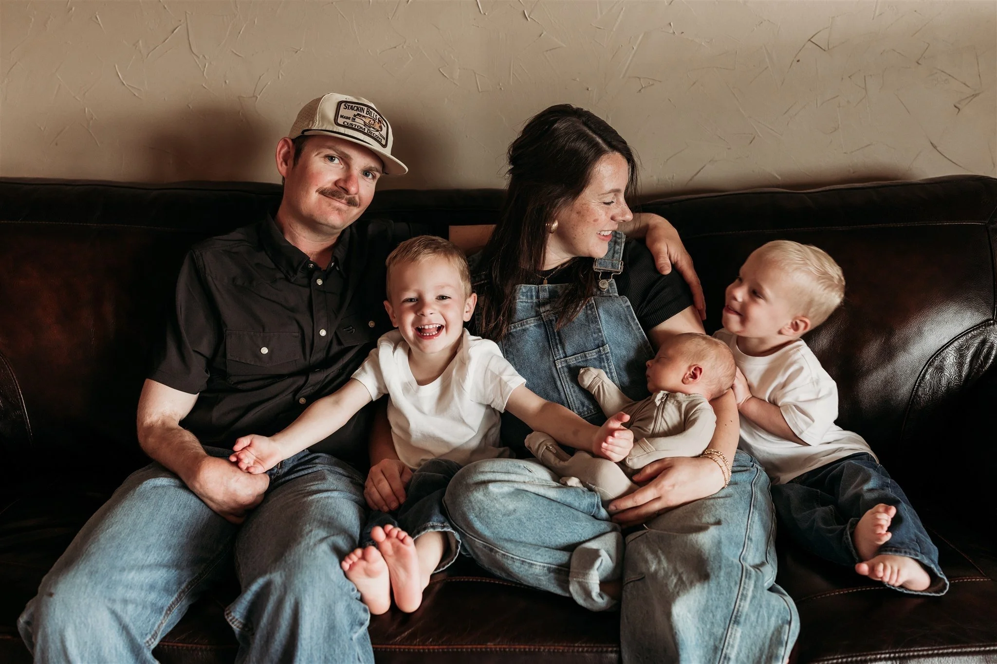 A family of five sitting on a dark brown leather couch, including a man wearing a baseball cap, woman with long dark hair holding a baby, and three young blonde boys, one of whom is sitting on the woman's lap.