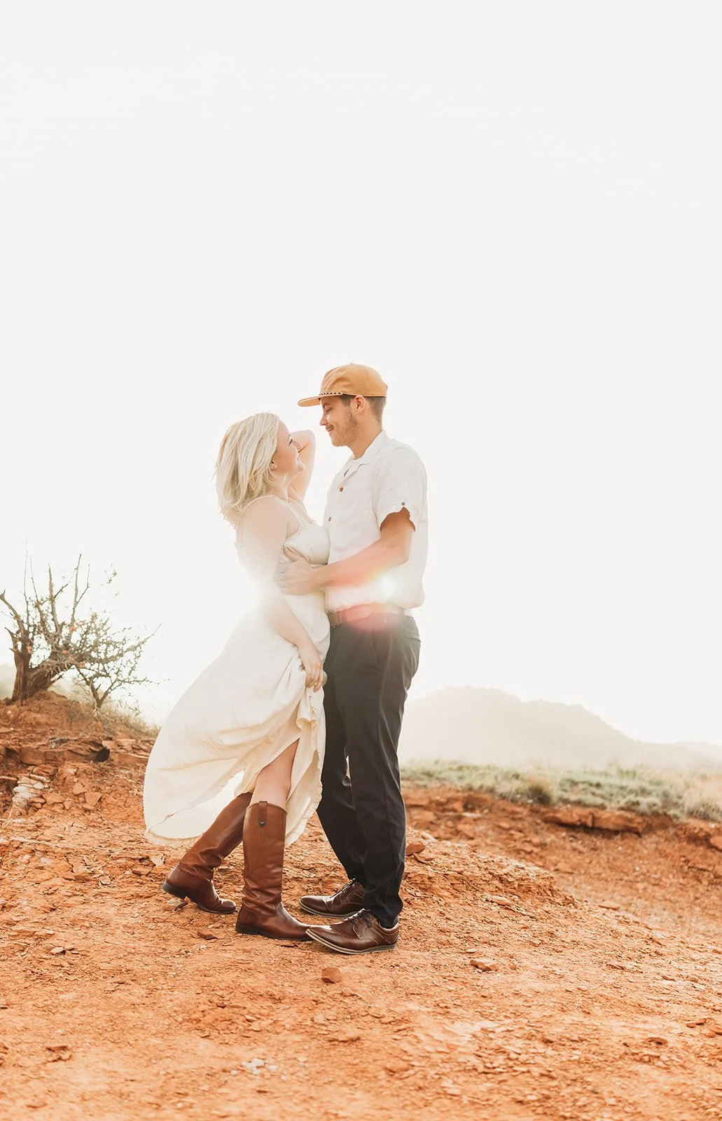 A couple standing close together outdoors on a dirt path with a barren tree and distant hills in the background, backlit by the sun, creating a warm glow around them. Photographed at Palo Duro Canyon.