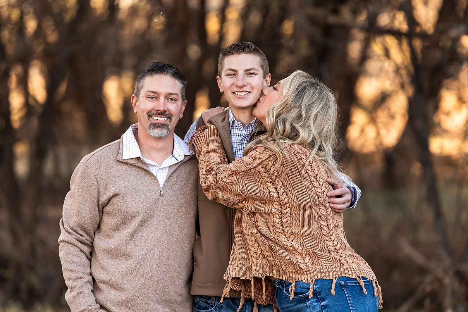 Family of three outside during autumn, with the woman kissing the young man on the cheek, and the man smiling at the camera.