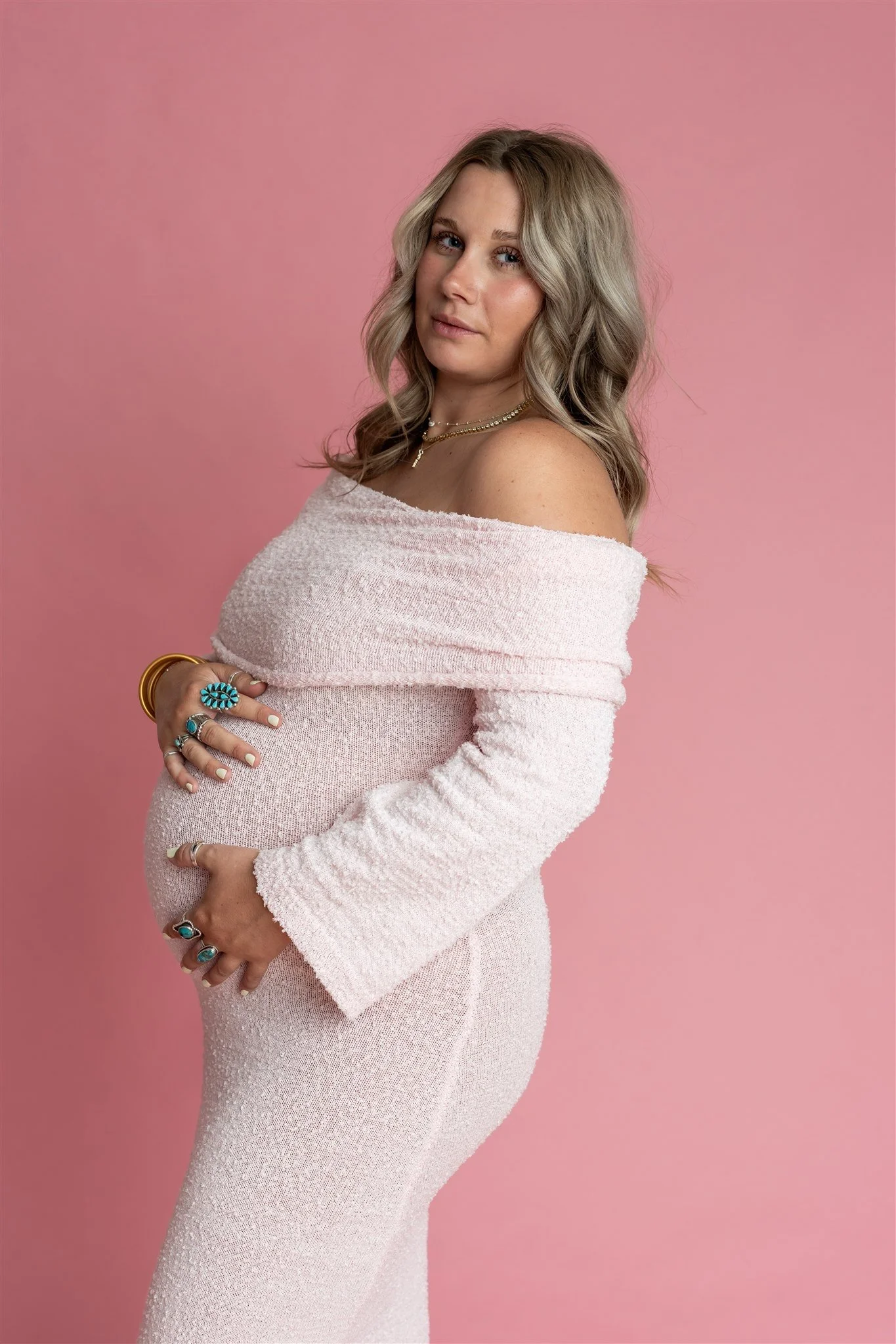 A pregnant woman with blond wavy hair wearing a light pink off-shoulder dress and multiple turquoise jewelry pieces stands against a pink background.