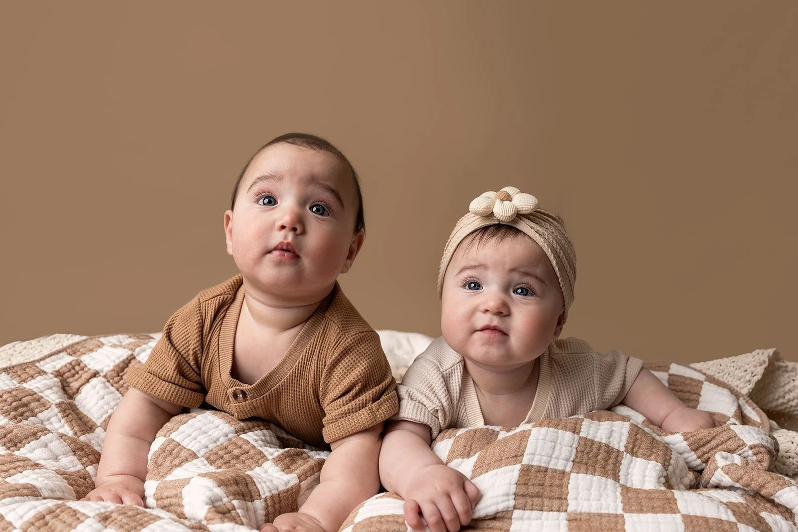 Two babies lying on a beige and white checkered blanket against a beige background. The baby on the left has short dark hair and is wearing a brown shirt. The baby on the right is wearing a beige headwrap with a flower decoration and a beige shirt.