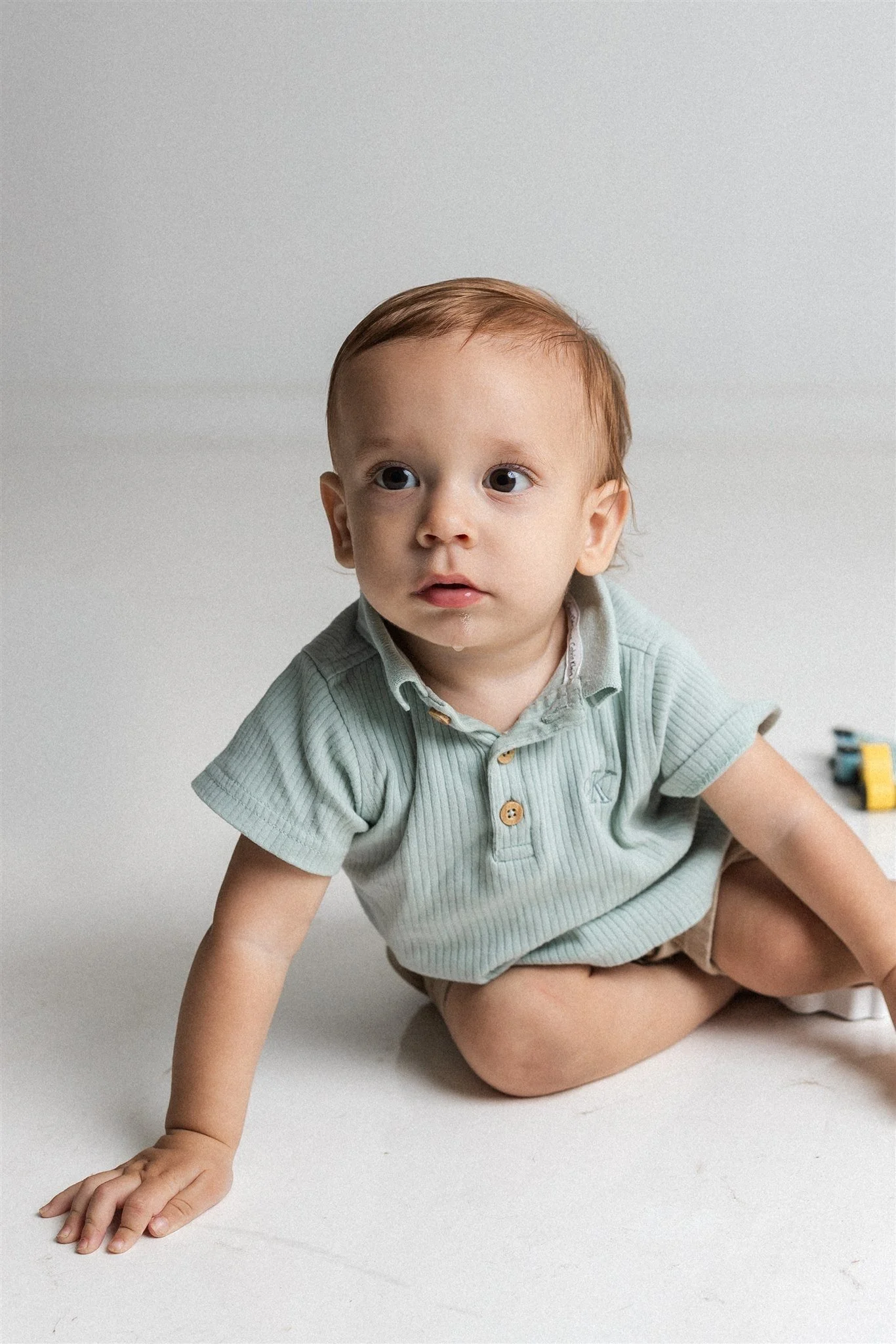 A young boy sitting on the floor, wearing a light green polo shirt and tan shorts, looking to the side with a curious expression.