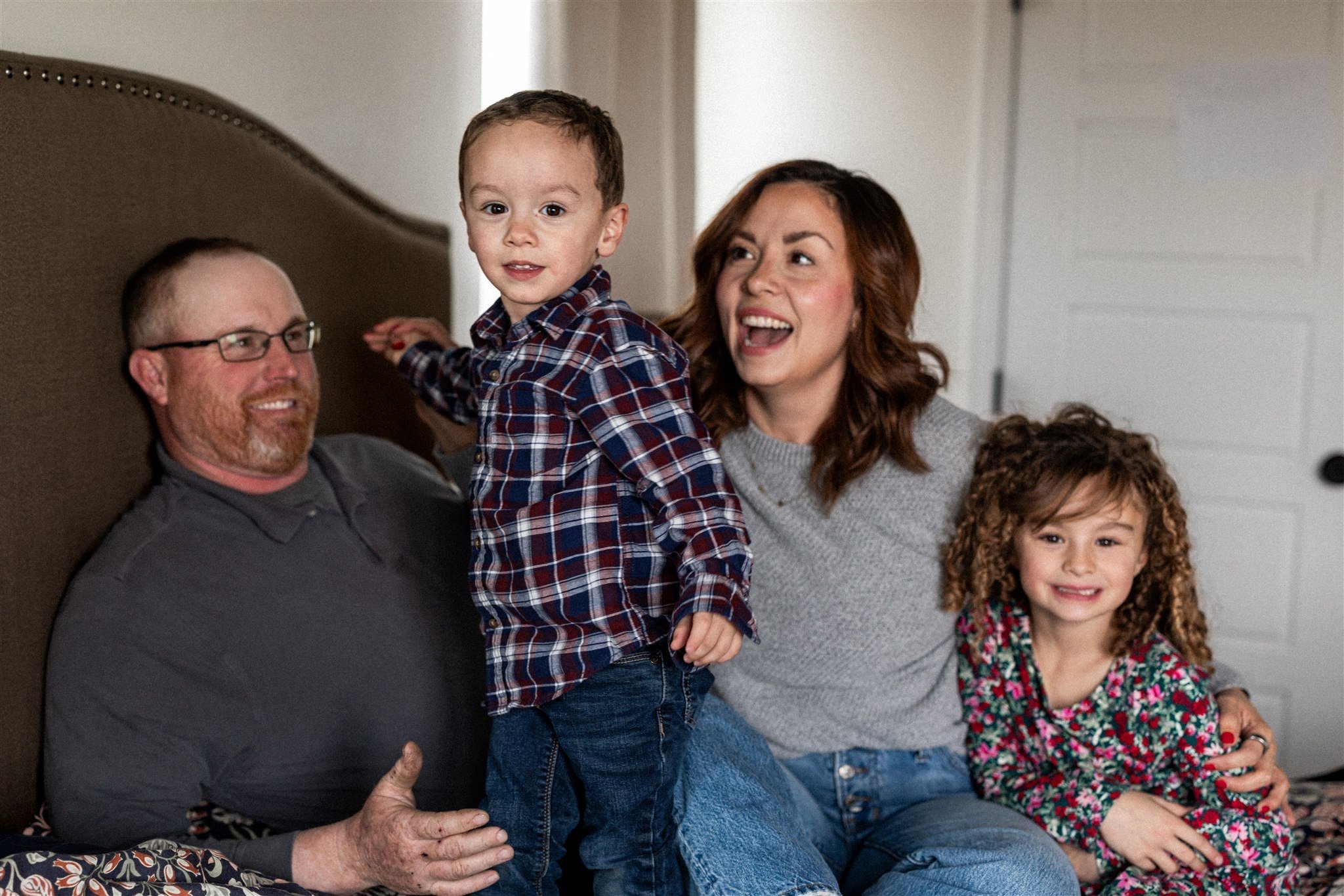 A happy family of four sitting on a bed, smiling and playing together. The father has a beard, glasses, and is wearing a gray shirt. The mother has shoulder-length wavy hair, wearing a gray sweater. The young boy is sitting on the father's lap, weari