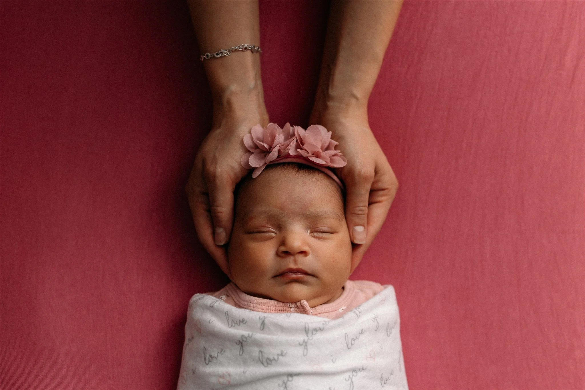 A newborn baby girl sleeping, wrapped in a blanket with 'love you' written on it, wearing a pink headband with three pink flowers, and being gently held by adult hands on a pink background.