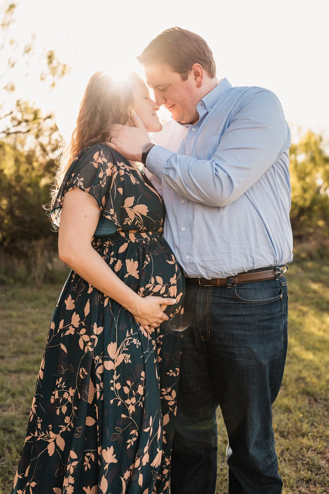 A maternity photo of a pregnant woman and her partner outdoors during sunset, with the man touching her face and holding her belly.