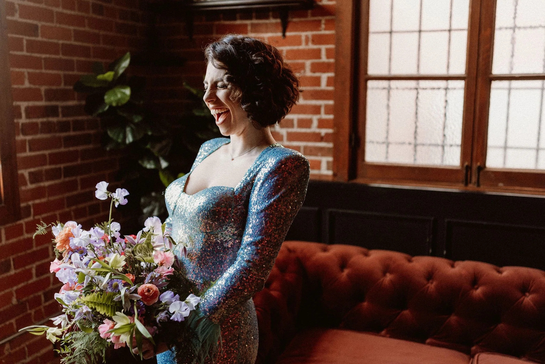 A modern bride in a sparkling blue sequin jumpsuit holds a vibrant bouquet with pink and purple flowers, standing in Carondelet House with exposed brick and large windows.