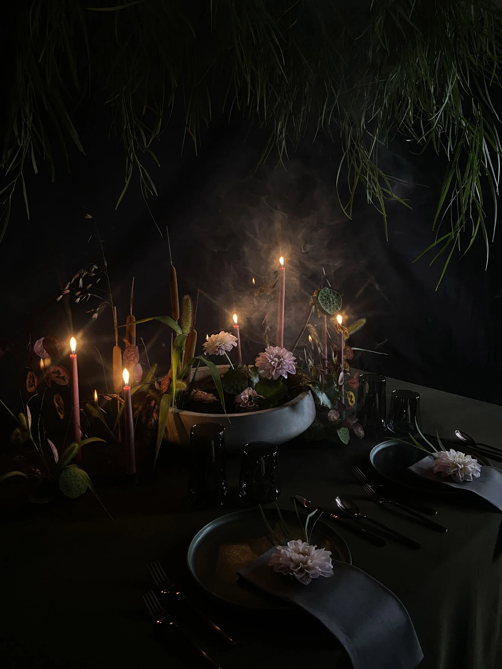 An enchanting dinner setting under a canopy of willow branches, illuminated by soft candlelight. The table features sleek black plates, complemented by deep grey linens and a centerpiece of  water lily inspired flowers and greenery, creating a myster