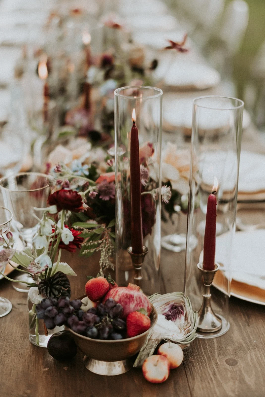Close-up view of a luxurious Dutch Masters inspired table setting featuring deep red candles in tall glass holders, a variety of fresh fruits like grapes and pomegranates, and a lush floral arrangements with dark dahlias and light blue tweedia, set a