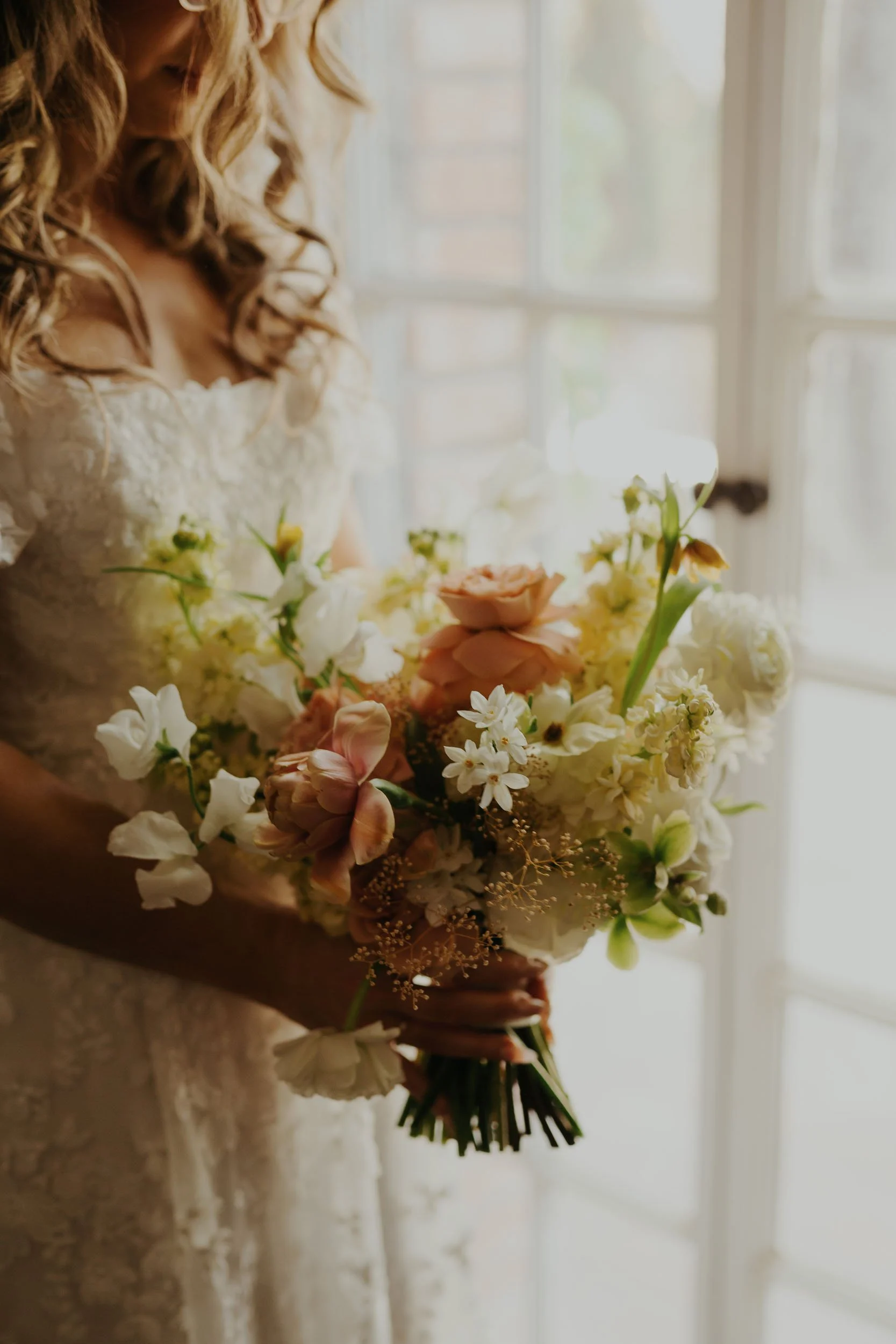 Close-up view of a bride holding a delicate wedding bouquet featuring soft peach roses, white sweet-peas, and green accents, illuminated by natural light near a window, enhancing the intricate lace details of her wedding gown.