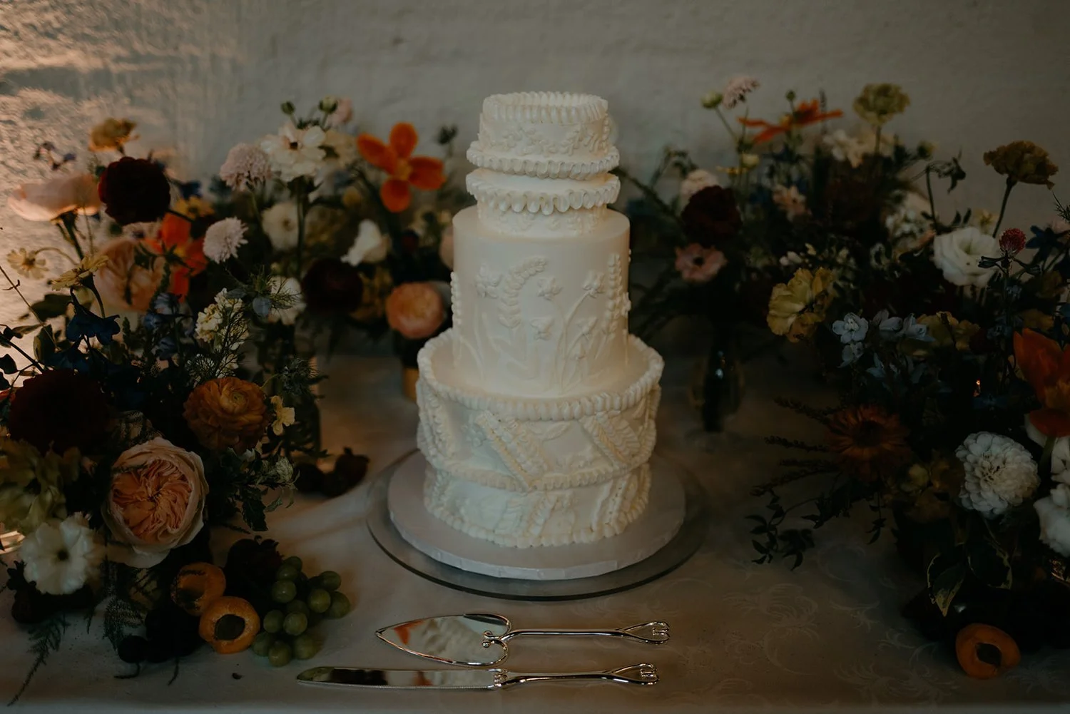 Exquisite three-tier wedding cake with intricate white icing designs, flanked by a lush arrangement of multicolored flowers and greenery, set against a textured white backdrop, creating a visually stunning centerpiece at a wedding reception
