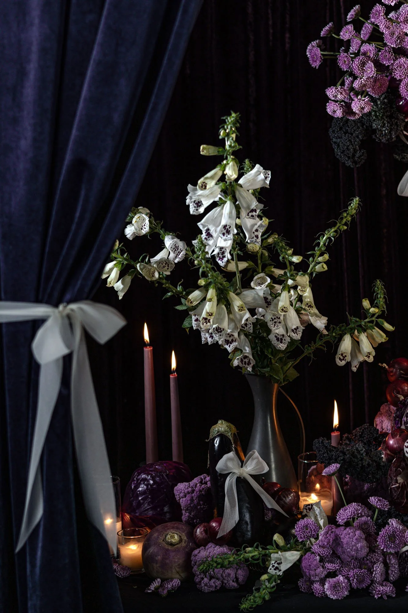 A captivating floral arrangement set against a dark velvet backdrop, featuring tall white foxgloves interspersed with dark purple accents, and surrounded by candles, creating a mysterious and Rembrandt inspired setting. This dramatic scene is further