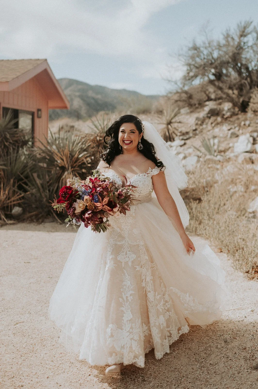 Bride in a flowing lace wedding gown holding a colorful bouquet of deep red, purple, and burgundy flowers, with a scenic mountain backdrop, captured in a desert setting