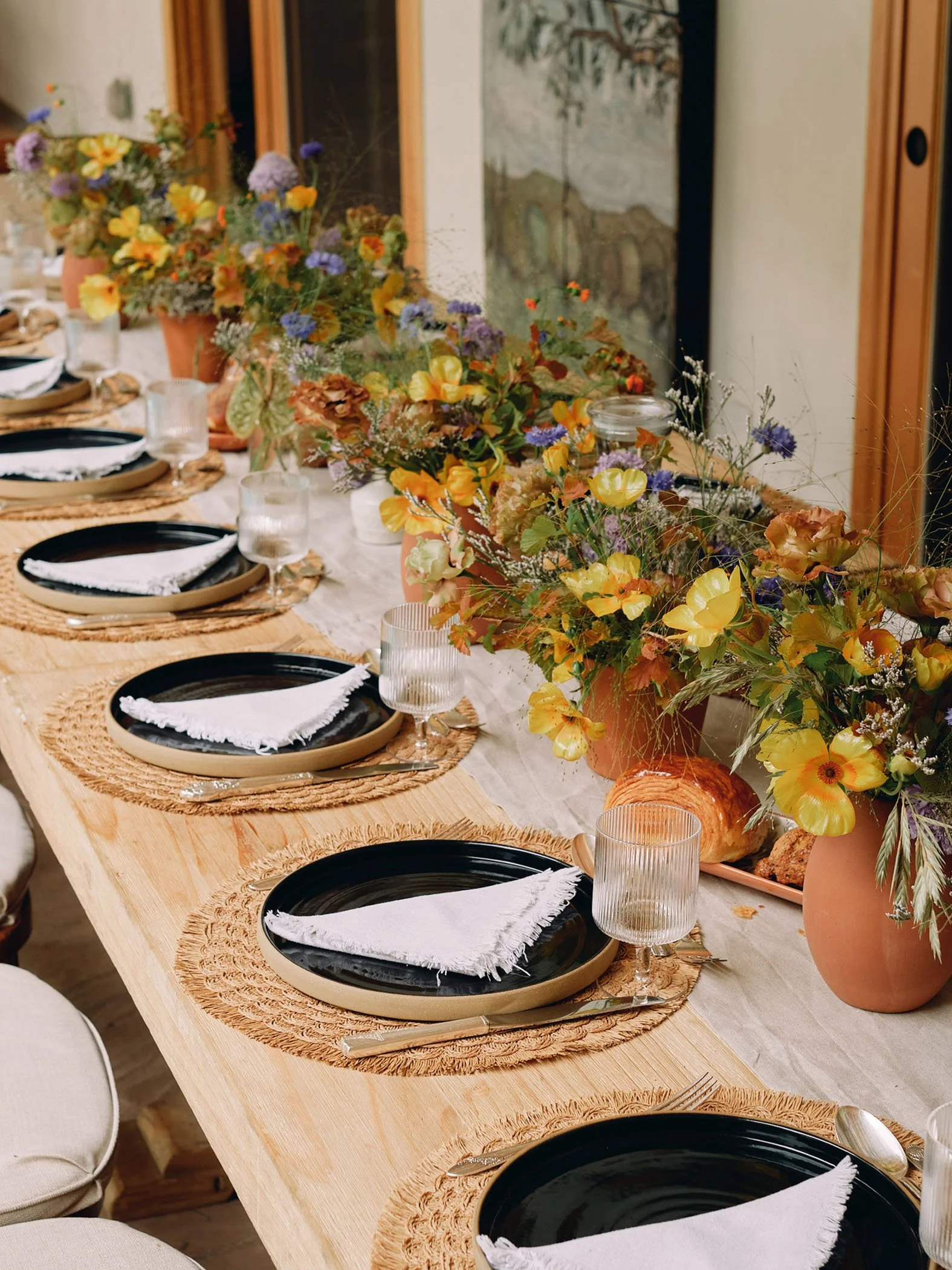 Long wooden table beautifully arranged for a garden event, adorned with vibrant flower arrangements in orange, yellow, and purple, set in terracotta pots. The table features rustic rattan chargers, sleek black plates, and soft white napkins, perfectl