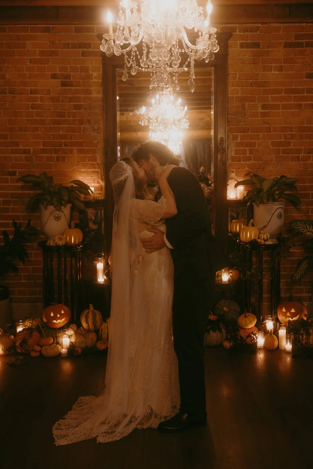 Romantic Halloween wedding moment featuring a couple sharing their first kiss surrounded by candlelight, pumpkins, and warm ambient decor in a moody indoor venue with brick walls and chandelier lighting.