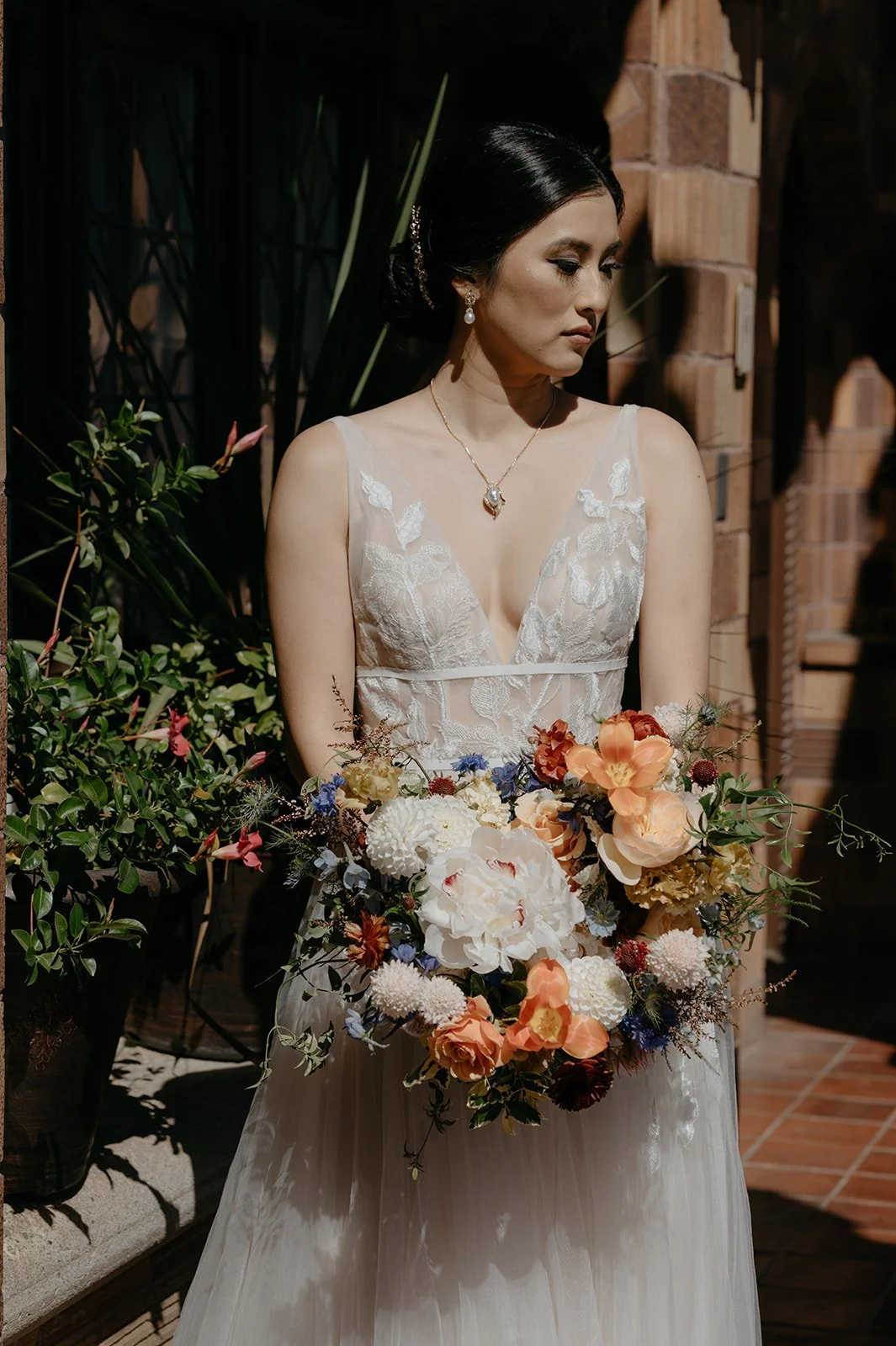 Elegant bride in a lace gown holds a luxurious Dutch Masters inspired bouquet with white peonies, blue cornflowers, and peach roses, set against a historic courtyard backdrop with lush greenery and architectural details at the Mission Inn in Riversid