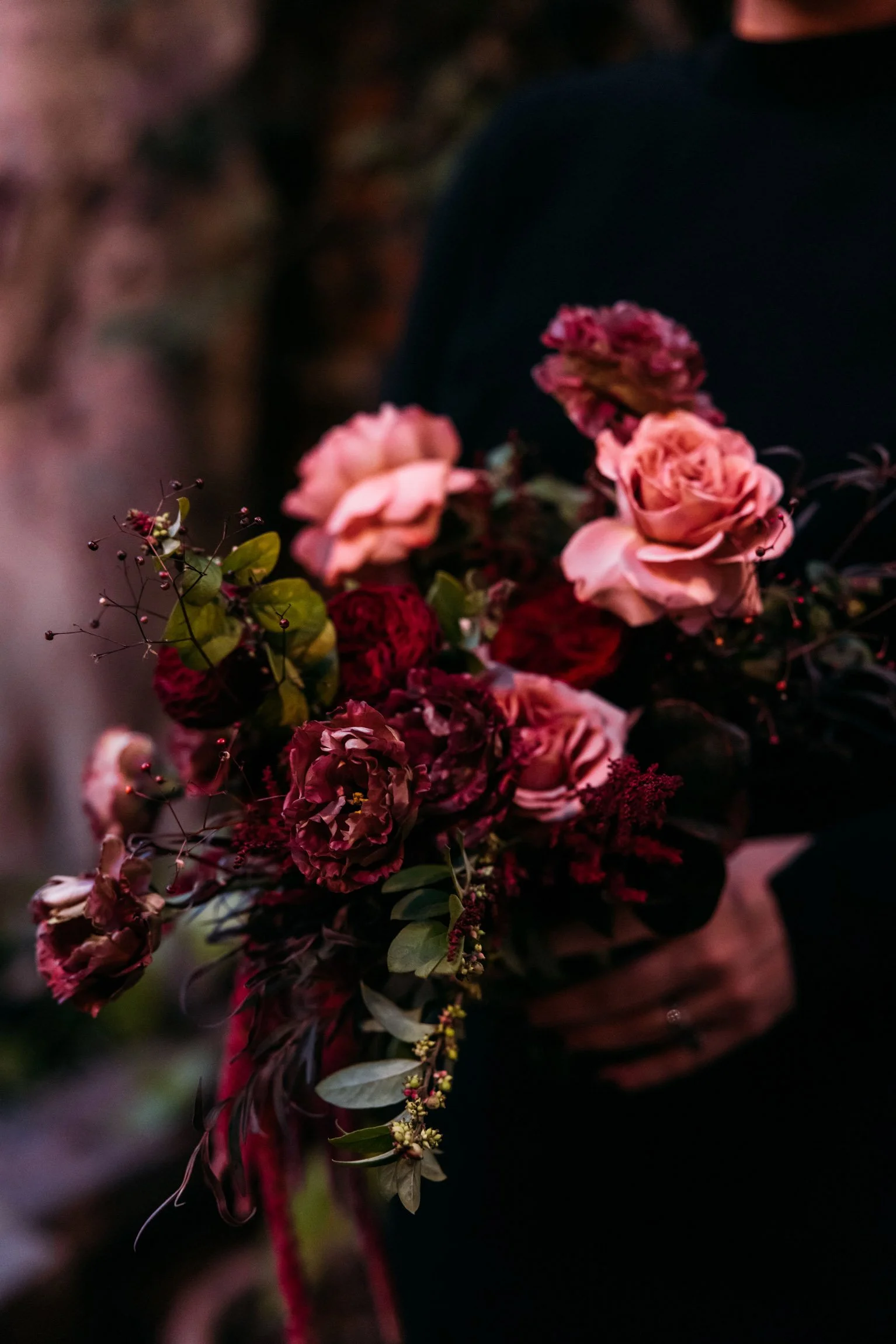 Close-up of a bride holding a lush autumnal bouquet with deep reds, dusty pinks, and hints of burgundy, surrounded by delicate greenery and textured accents, capturing the essence of the romantic fall season.