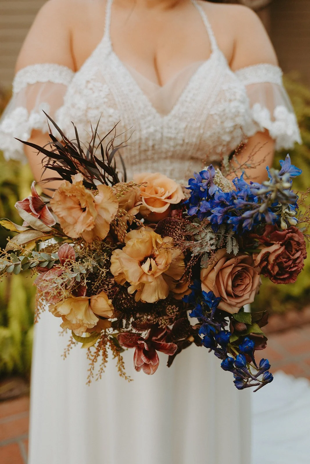 Vivid close-up of a bride holding a striking autumn wedding bouquet, featuring deep blue delphiniums, golden toned roses and unique foliage accents, set against a sparkling bead-embellished wedding gown.