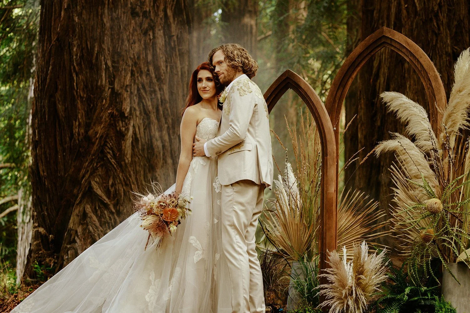 Intimate Redwoods wedding scene with wooden cathedral arches adorned with grasses, palm leaves and dried florals, surrounded by towering trees, enhancing the natural and serene ambiance.