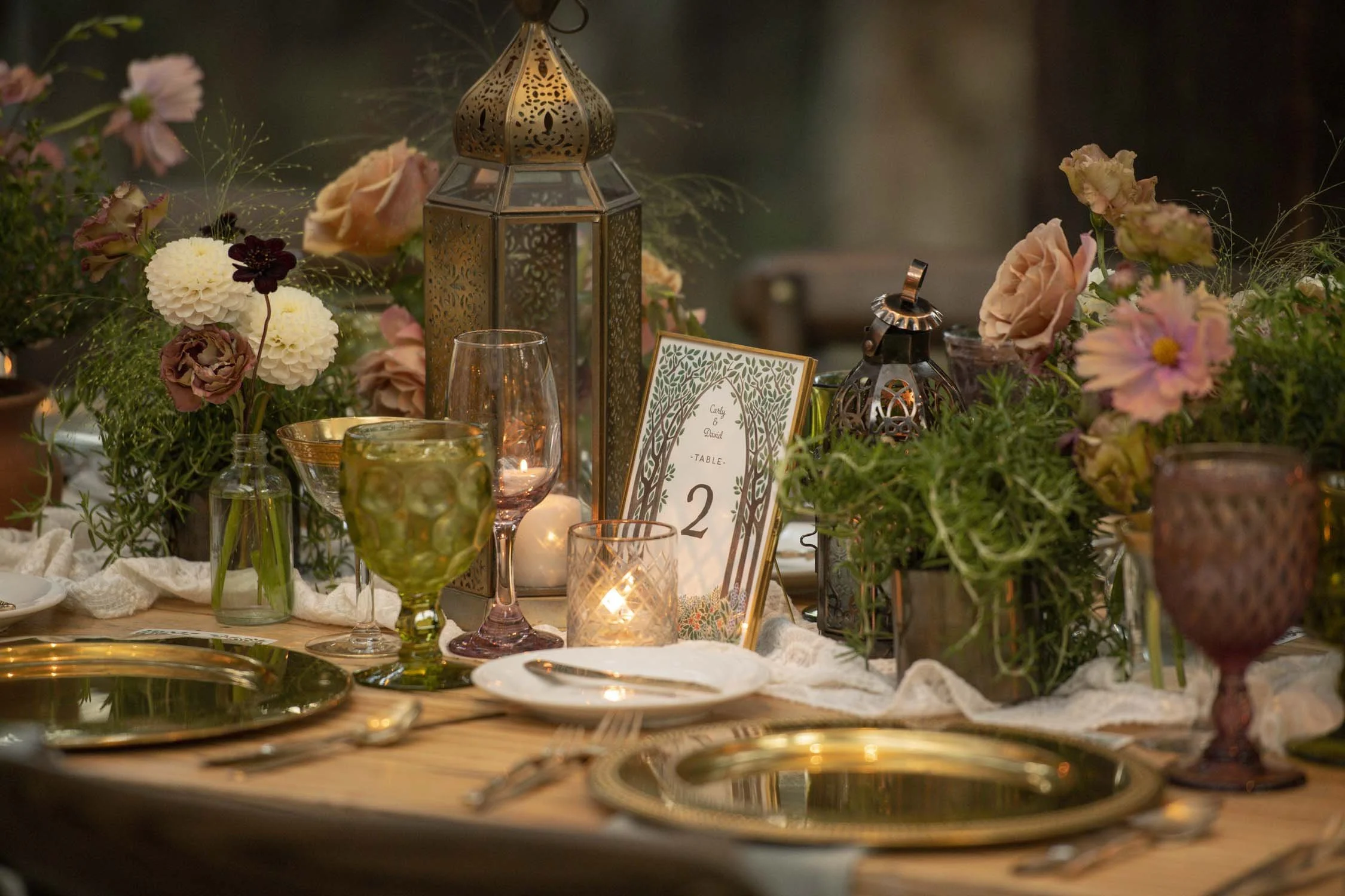An intimate wedding table setting showcasing an organic and elegant look, with lush floral centerpieces of dusty pink cosmos, cream dahlias, herbs, and potted plants complemented by ornate golden lanterns and colored glassware, all set against a back