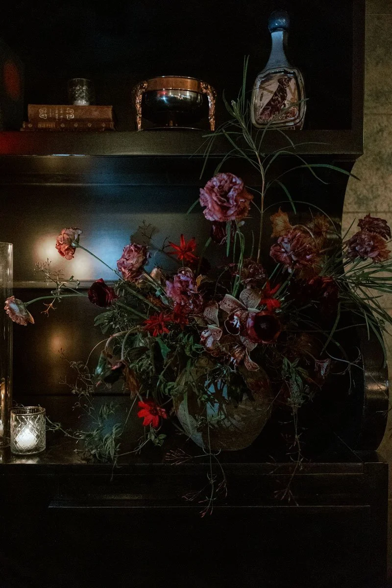 A dramatic floral arrangement at Hotel Per La, featuring rich, dark hues of red and brown blooms with delicate textures, set against a backdrop of a dark wooden shelf, evoking a sense of mystery and old-world charm.