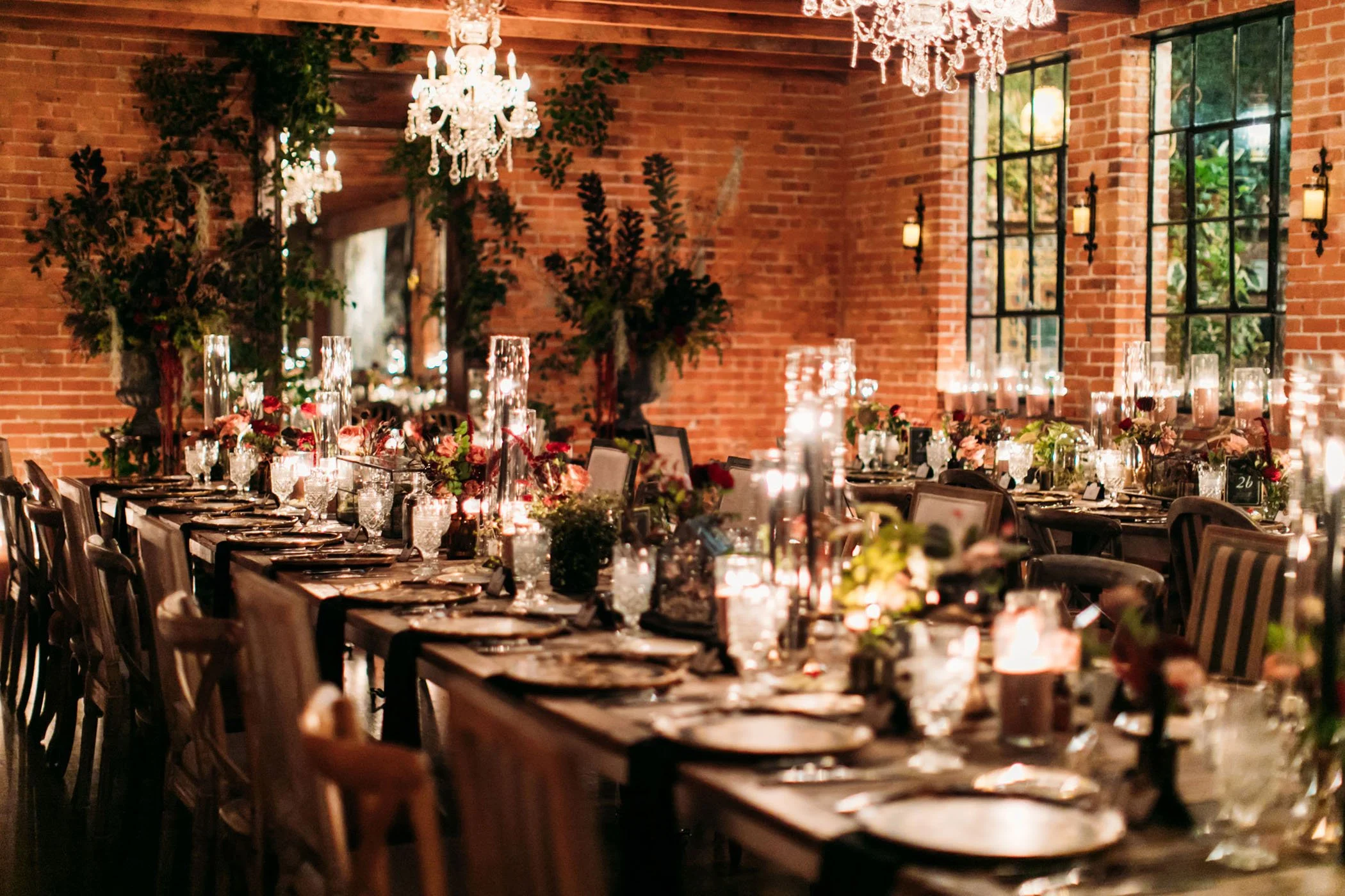 A beautifully decorated wedding reception table set in the Carondelet House venue with exposed brick walls, featuring candlelit ambiance, lush floral centerpieces in deep reds and greens, crystal chandeliers, and a reflective table setting that enhan